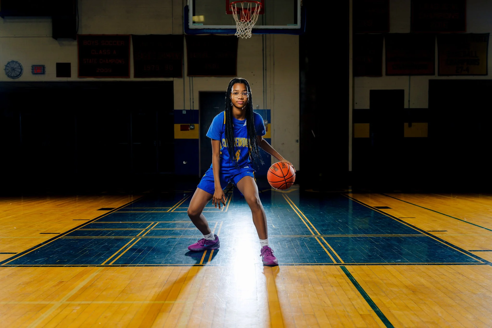 A young girl in a blue basketball uniform standing on a basketball court, holding a basketball, preparing to dribble or pass, in a gymnasium with banners on the wall.