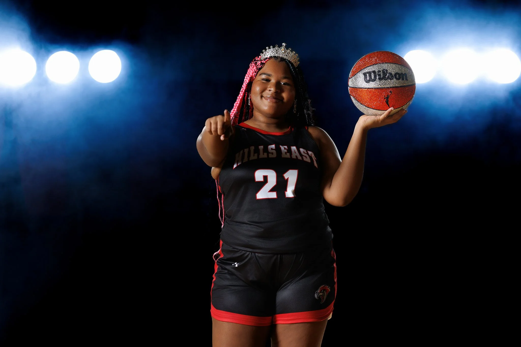 A young girl wearing a black and red basketball uniform with 'Hills East' and the number 21, holding a glittery basketball, wearing a silver tiara and smiling, standing in front of bright lights.