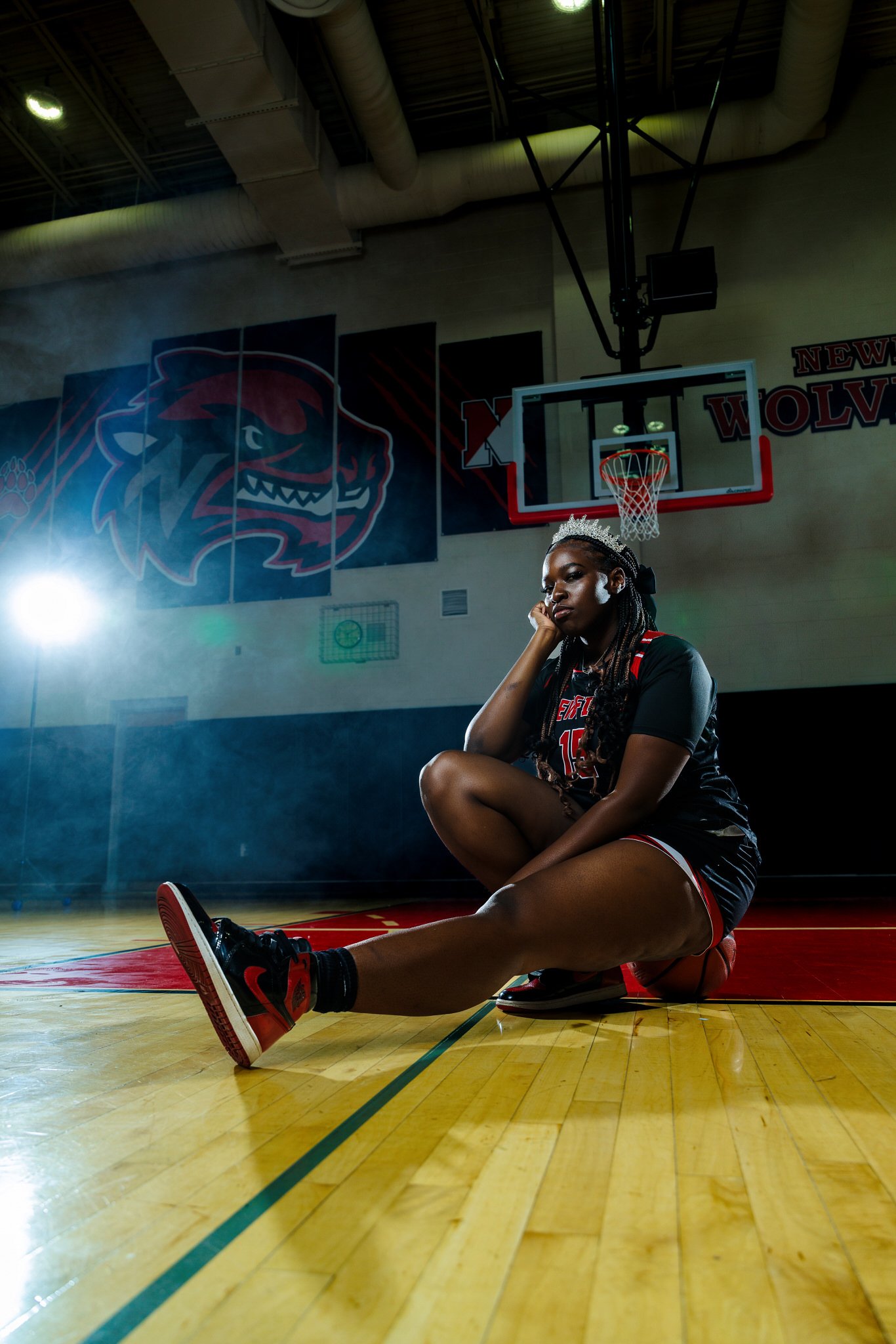 A female basketball player sitting on the court in a gym, wearing a black uniform with red and white accents, a tiara, and sneakers, looking contemplative.