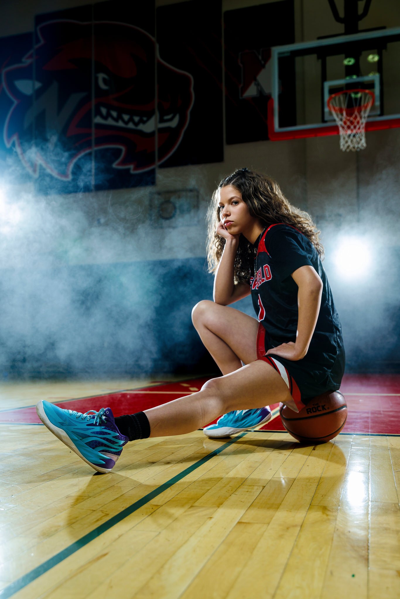 A young female basketball player sitting on the court with a basketball under her hand, looking serious, in a gym with a large cat logo on the wall and a basketball hoop in the background.