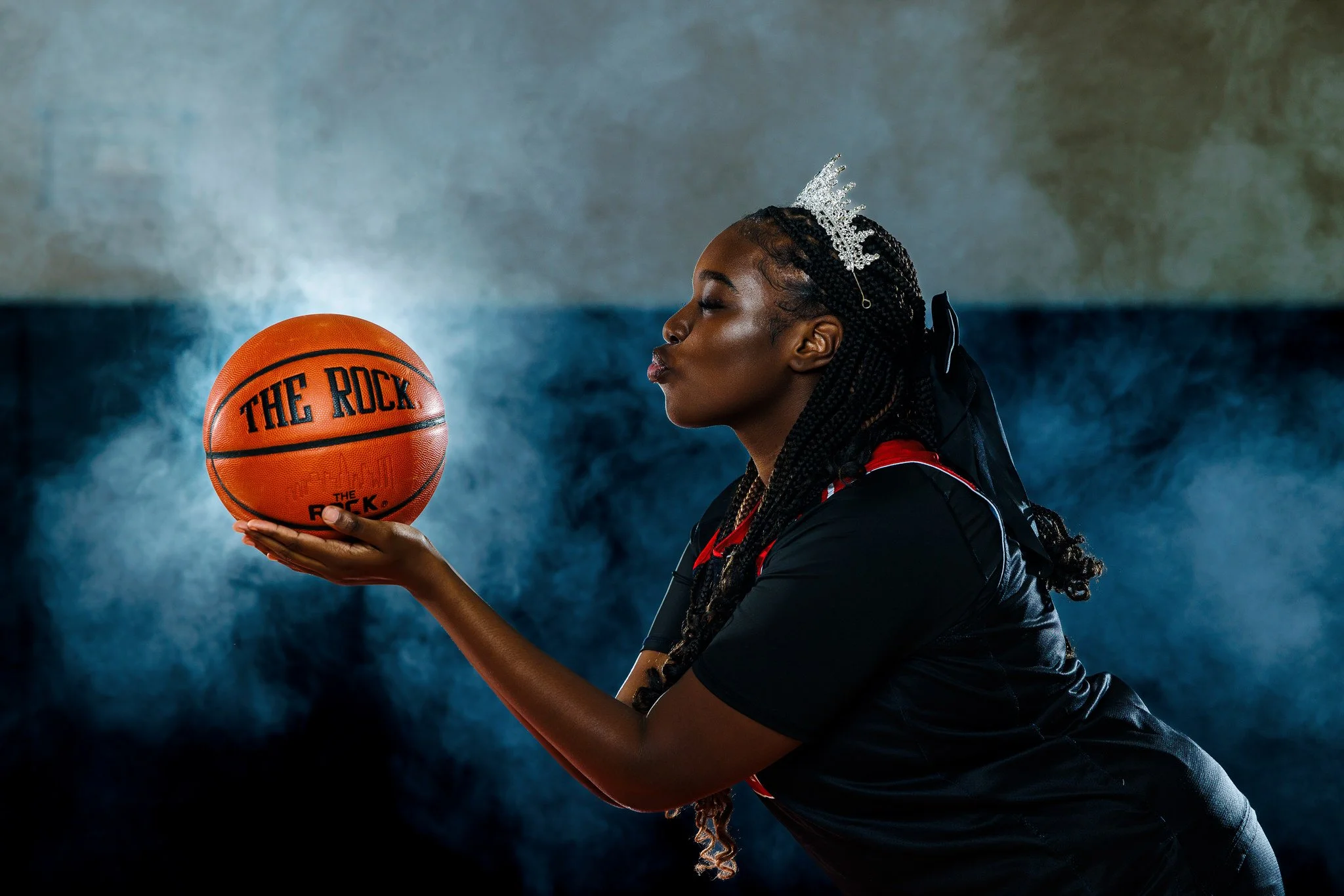 A woman holding a basketball labeled 'THE ROCK' with a serious expression, wearing a black sports outfit and a small crown hairstyle, standing in a gym with a blue and gray background.