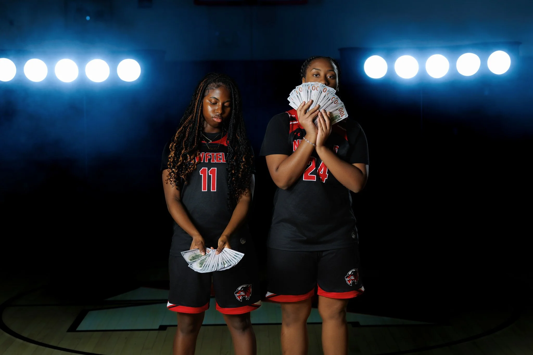 Two female basketball players in black uniforms with red accents and numbers 11 and 24, standing in a dark gymnasium with bright lights overhead, holding fanned-out stacks of cash.