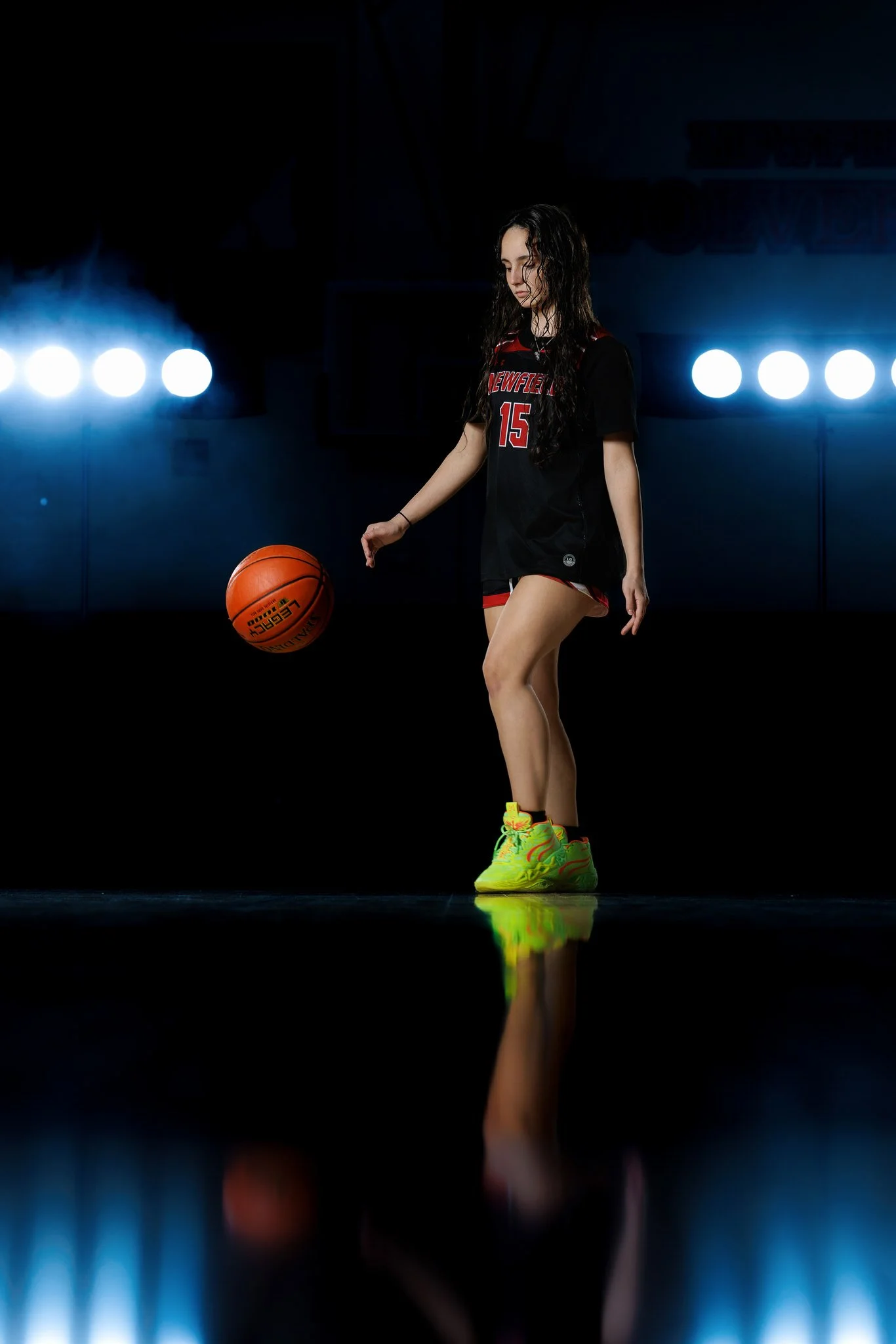 A young female basketball player with long curly hair, wearing a black and red uniform with the number 15, is standing on a dark basketball court, dribbling an orange basketball. Bright lights are shining behind her, creating visible reflections on t
