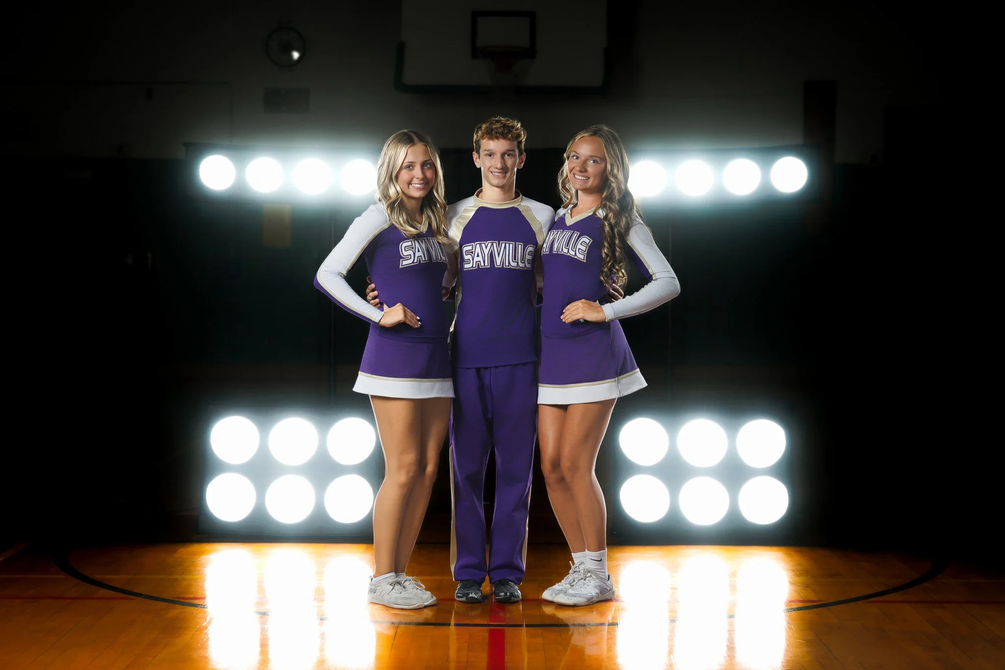 Three cheerleaders in purple and white outfits standing on a basketball court with bright stage lights behind them.