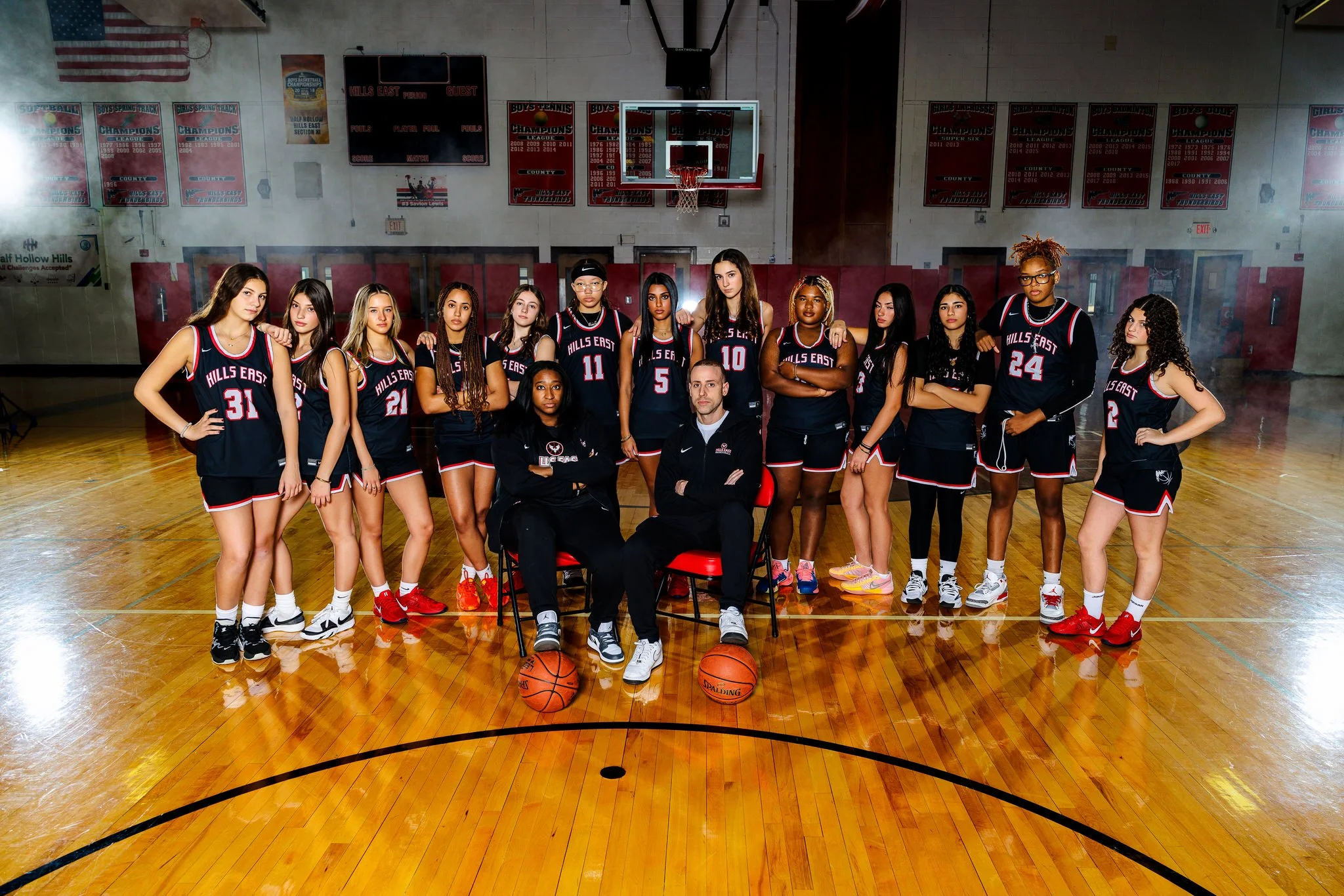 A girls' basketball team in black uniforms with red and white accents, posing on a gymnasium basketball court with their coaches. The team has 14 players standing and sitting in front of a basketball hoop, with basketballs on the floor. The gym has b