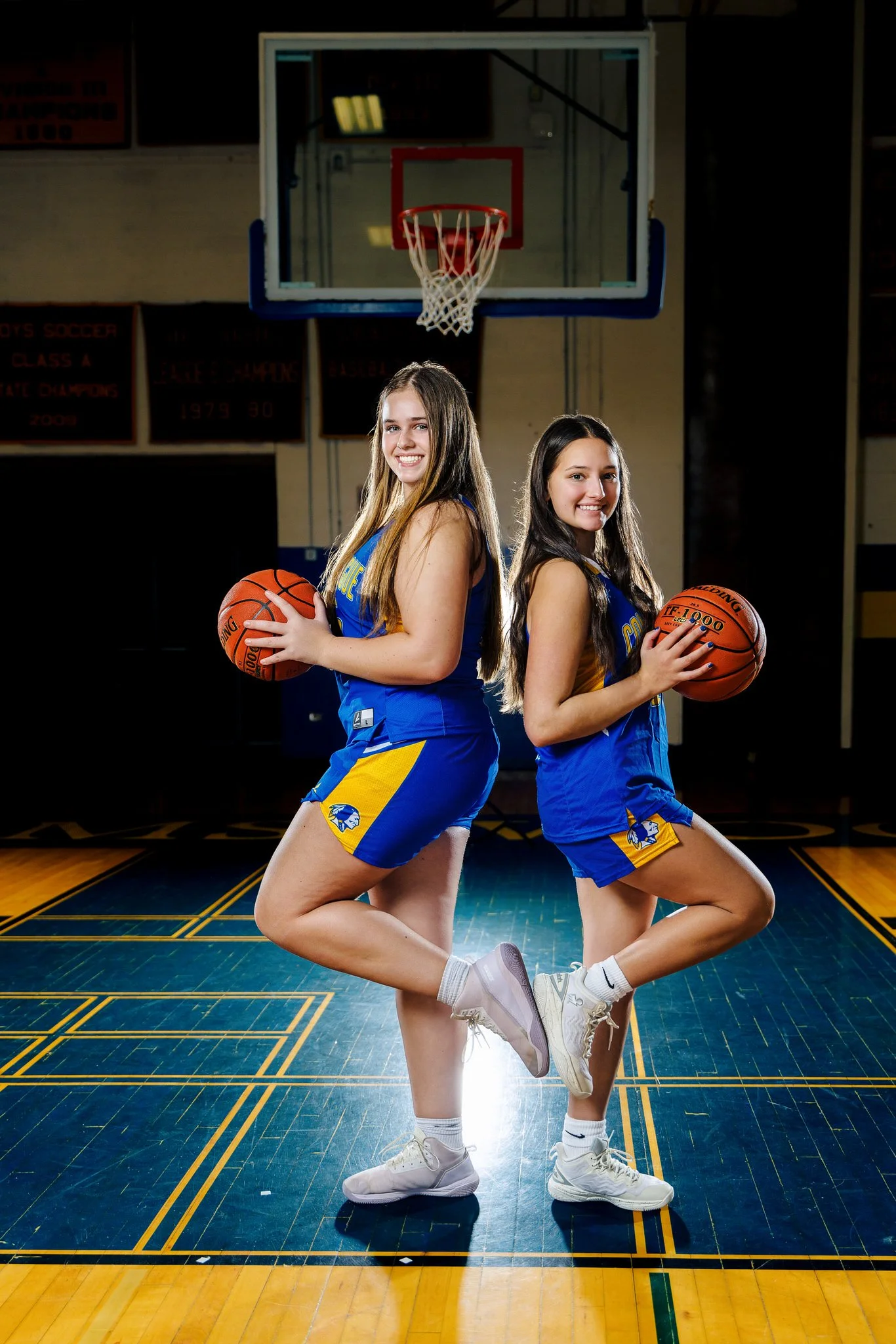 Two female basketball players in matching blue and yellow uniforms standing back-to-back on a basketball court, each holding a basketball, with a hoop overhead.