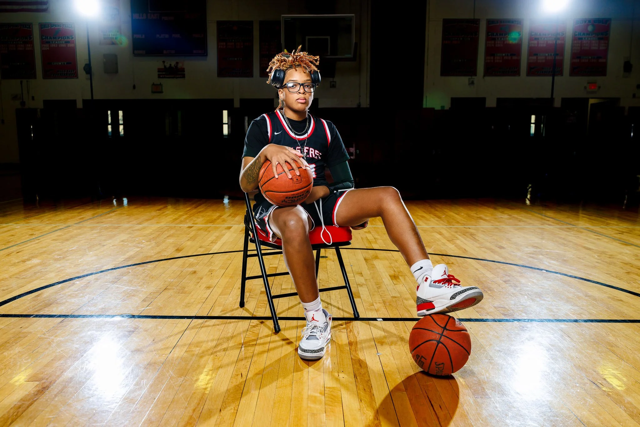 A young female basketball player sitting on a chair on a basketball court, holding a basketball with a second ball on the floor in front of her, wearing a black and red basketball uniform, headphones, glasses, and white sneakers.