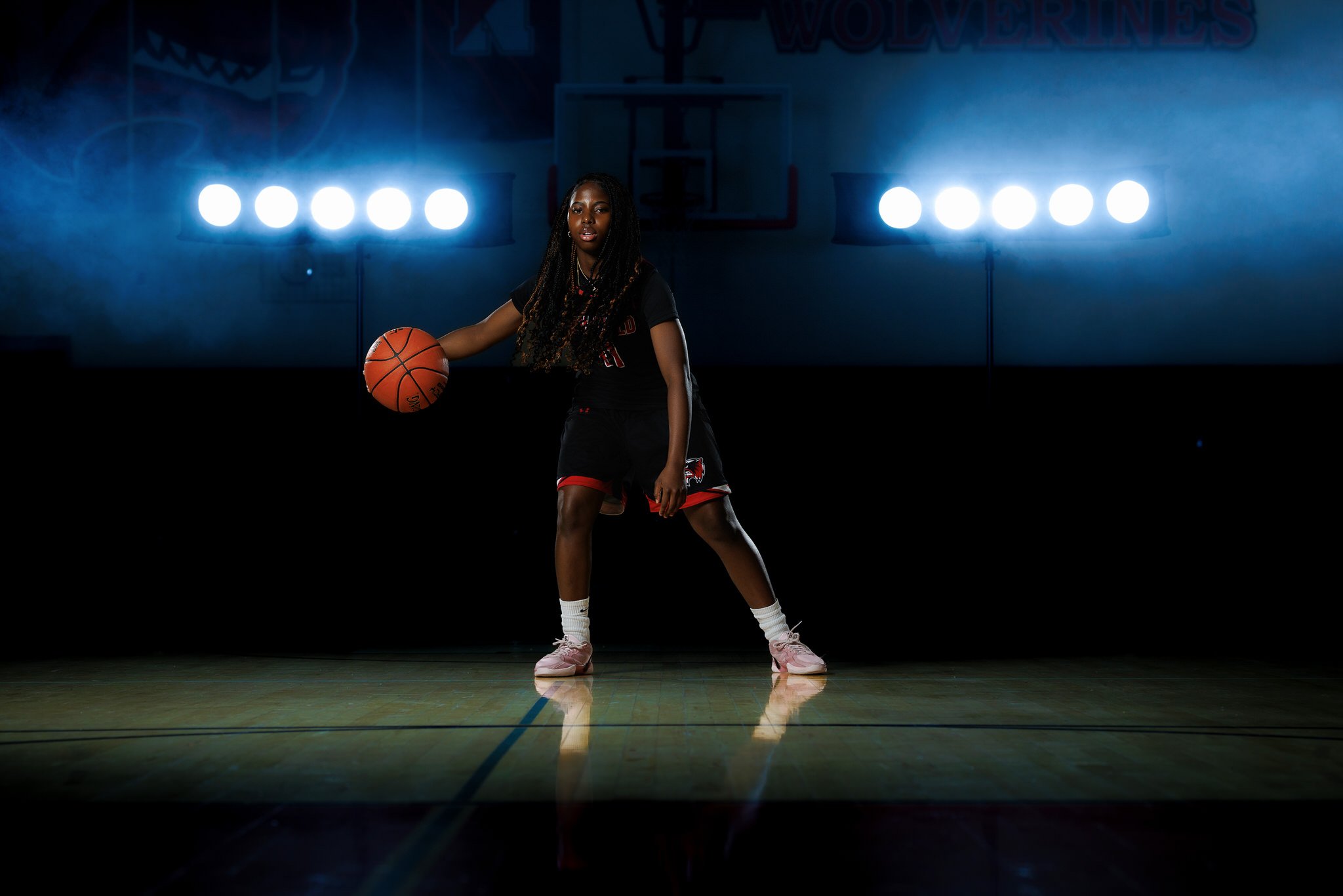 A female basketball player standing on a court with a basketball, in a dark gymnasium with bright lights behind her.