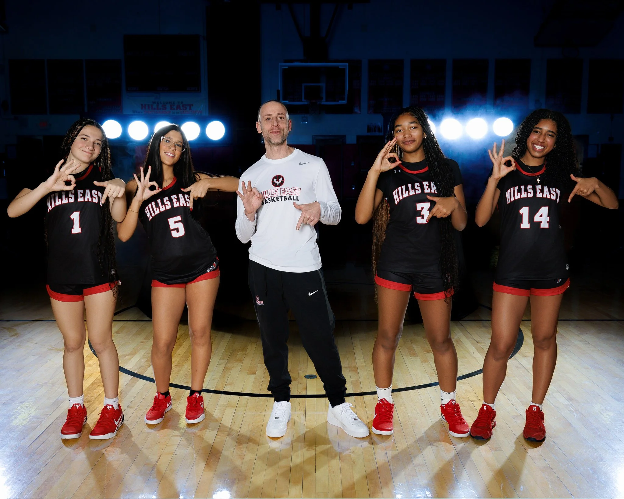 A volleyball coach and four female volleyball players standing on a gymnasium court, posing for a team photo. All are making hand gestures and smiling, with team uniforms that say 'HILLS EAST' and numbers.