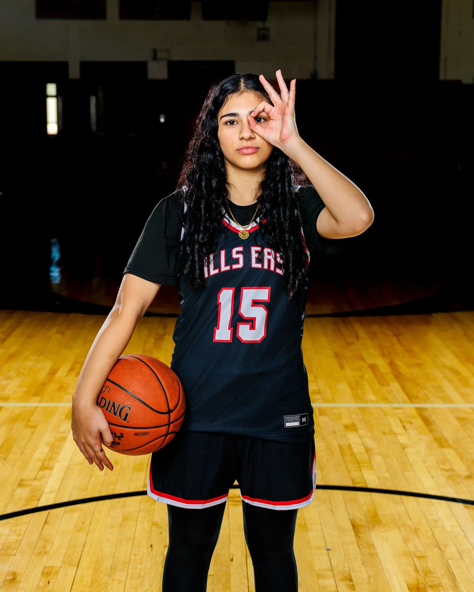 Young woman in a basketball uniform holding a basketball on an indoor court, making a gesture with her right hand around her eye, viewed from front.