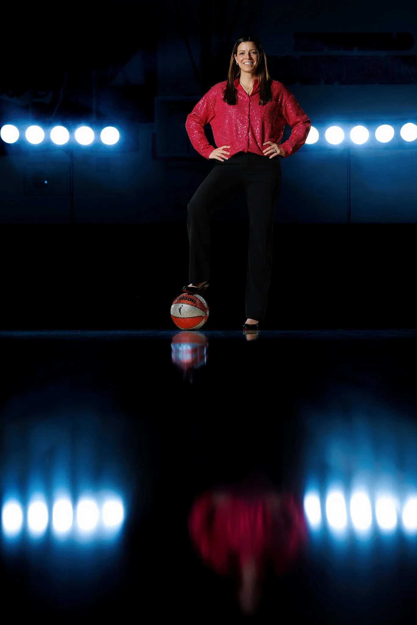 A woman in a red sequined shirt and black pants standing with one foot on a basketball, smiling in a dark setting with bright lights behind her.