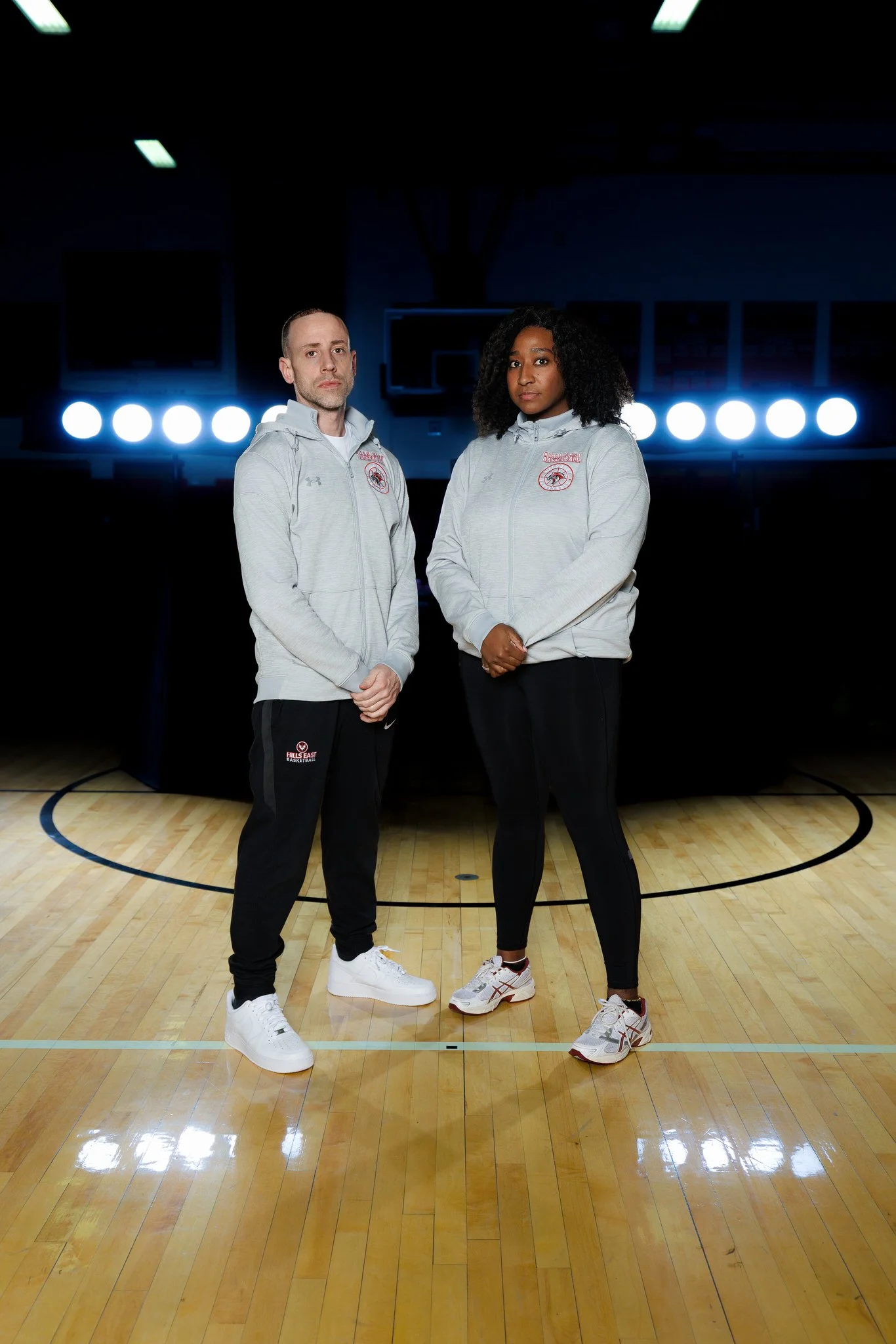 Two basketball players standing on a court, facing the camera, with bright lights and basketball hoops in the background.