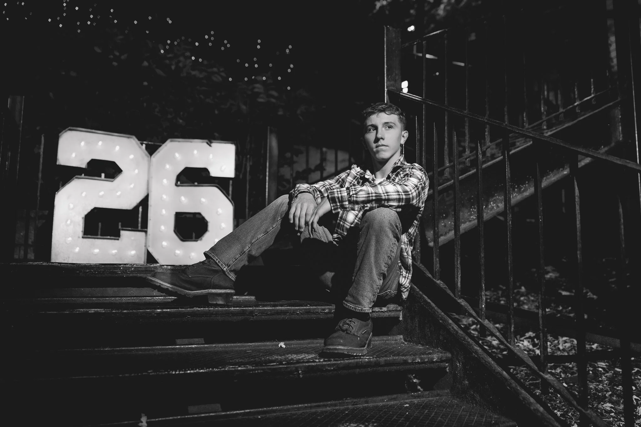 A young boy sits on dark outdoor steps at night, wearing a plaid shirt and jeans, with a large illuminated number 26 sign behind him.