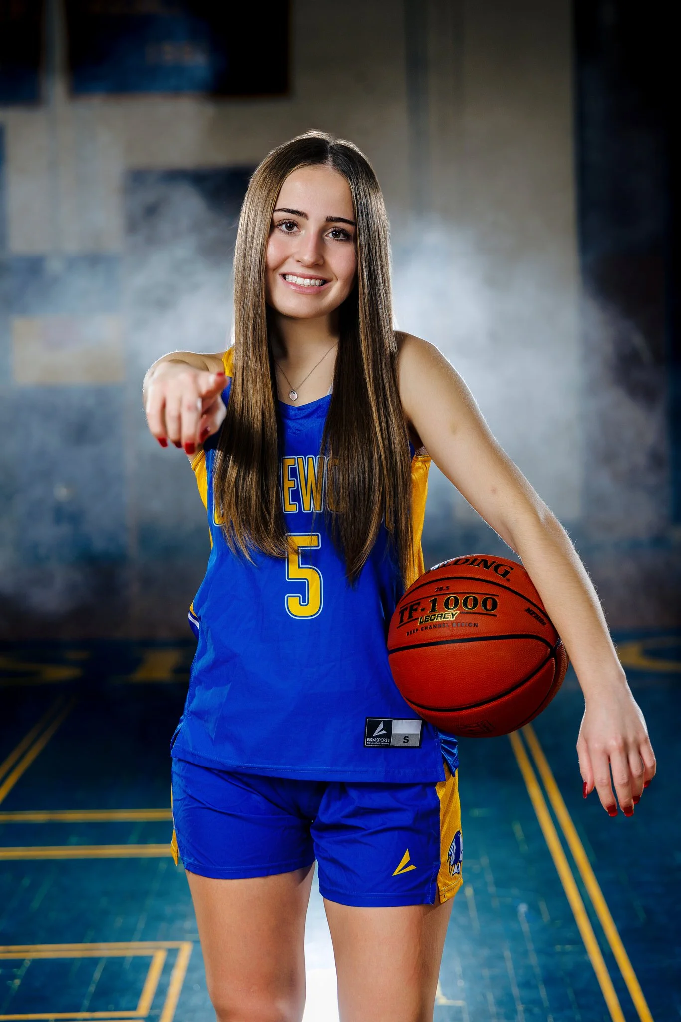 A young female basketball player in a blue and yellow uniform holding a basketball and pointing towards the camera inside a gym.