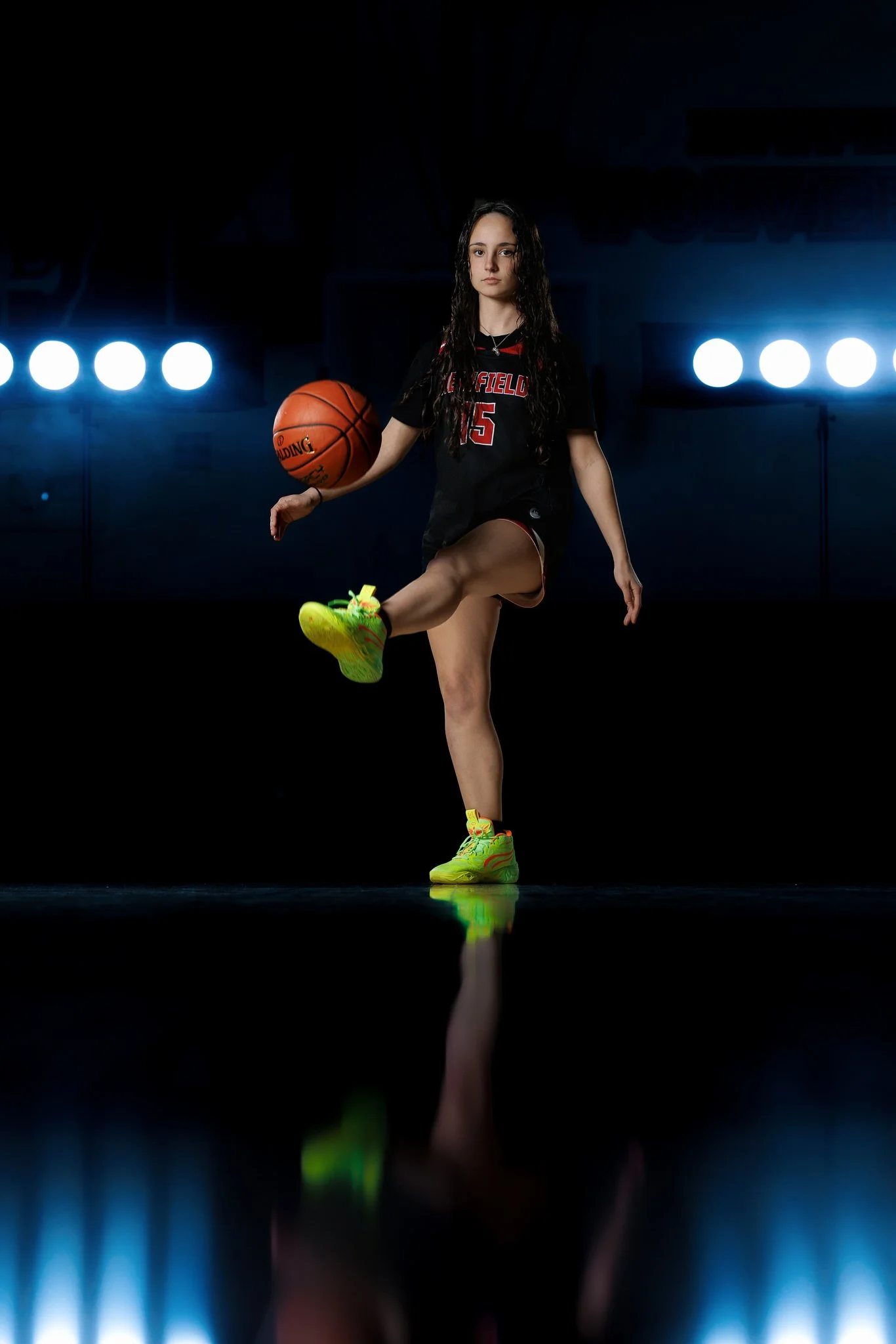 A young female high school basketball player in a black uniform with red accents practicing in a dimly lit gym, holding a basketball with one hand, one leg extended, and wearing neon yellow-green basketball shoes.