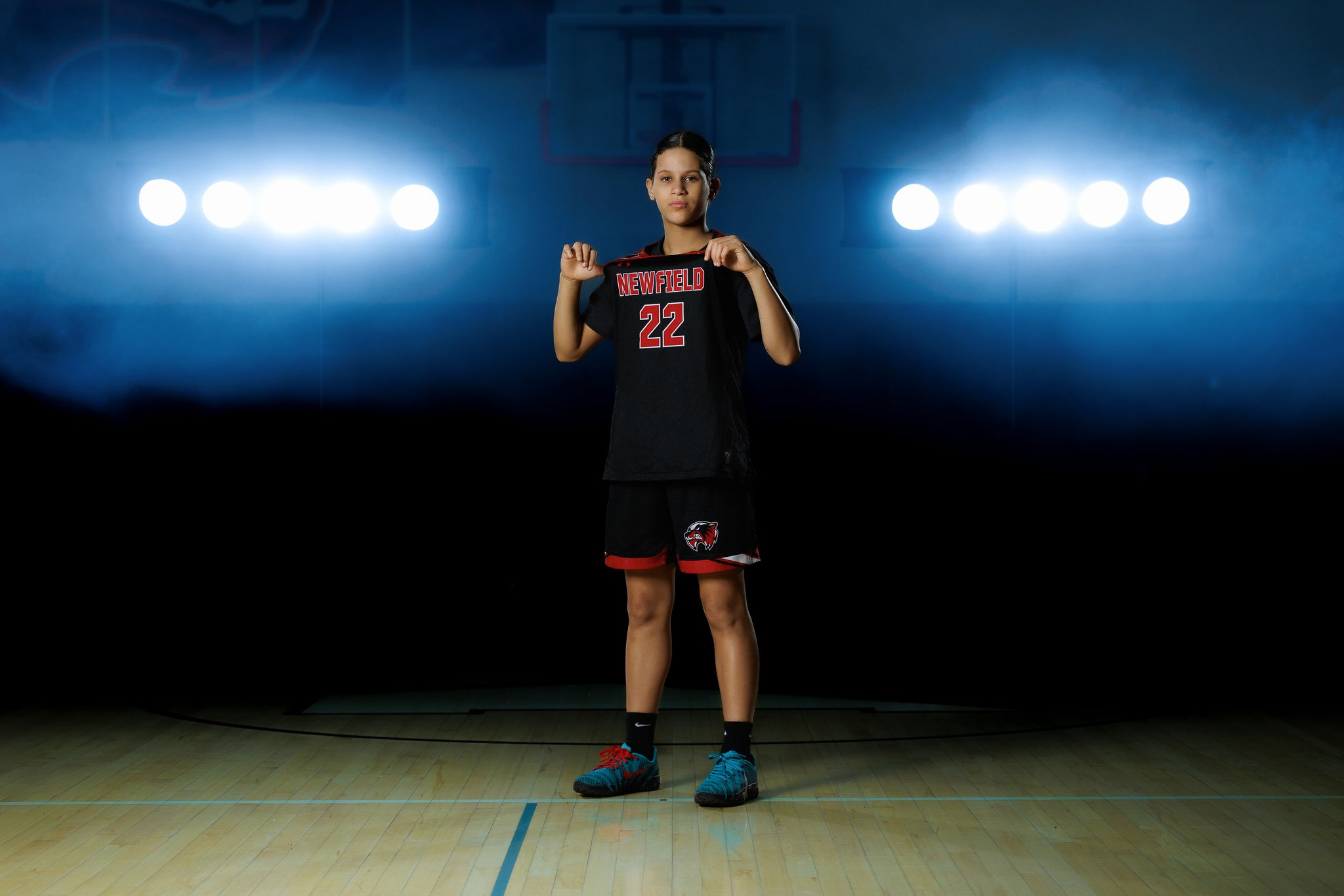 A young female volleyball player stands on an indoor court with bright lights overhead, wearing a black and red sports uniform with the number 22 and the word 'Newfield' on her jersey.