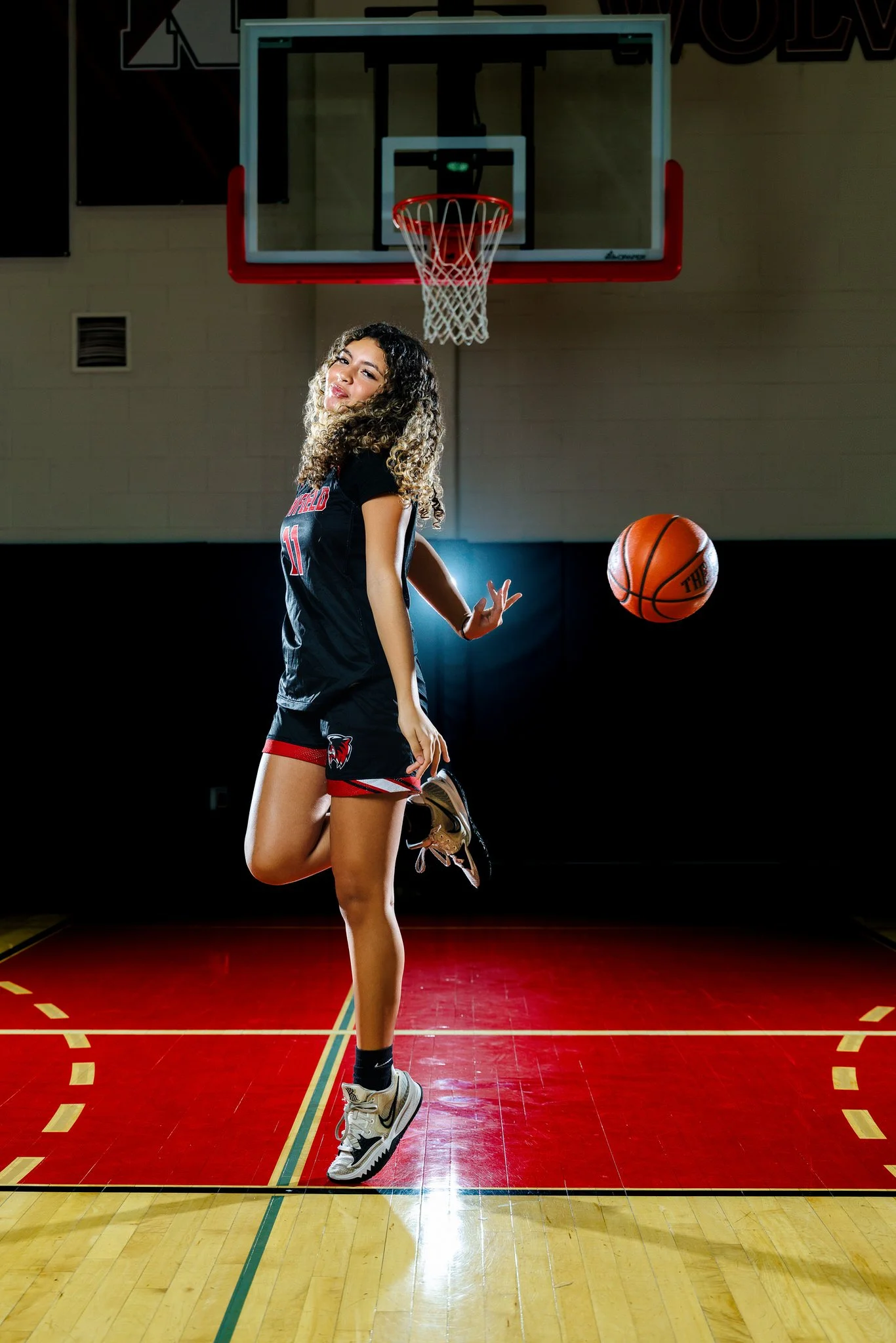 A young woman in a basketball uniform is standing on a basketball court in a gymnasium, near the basketball hoop, and is mid-move as she appears to be about to shoot or pass a basketball.