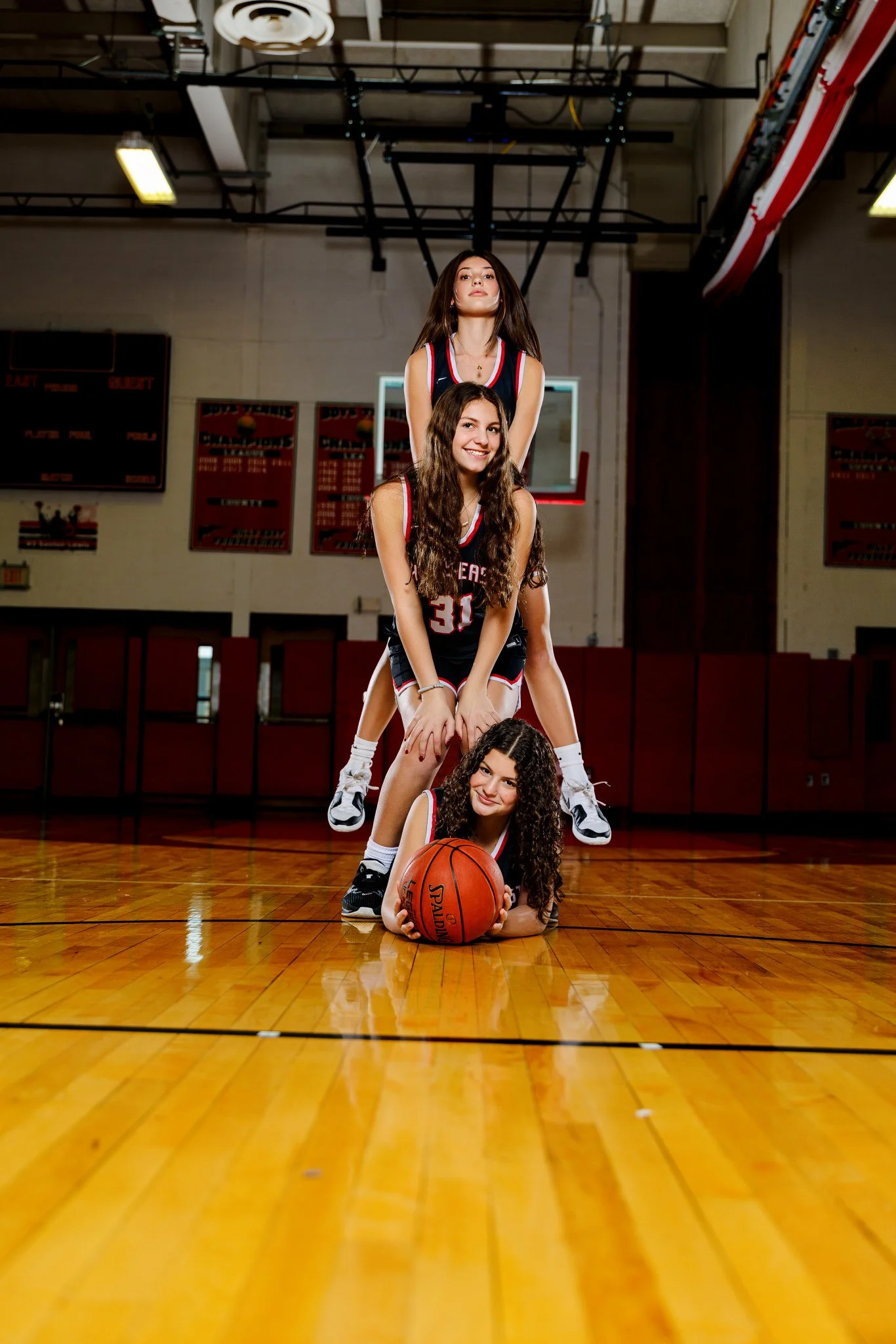 Four female basketball players stacked in a gymnasium on a basketball court, with a basketball at the front, all smiling and wearing black and red basketball uniforms.