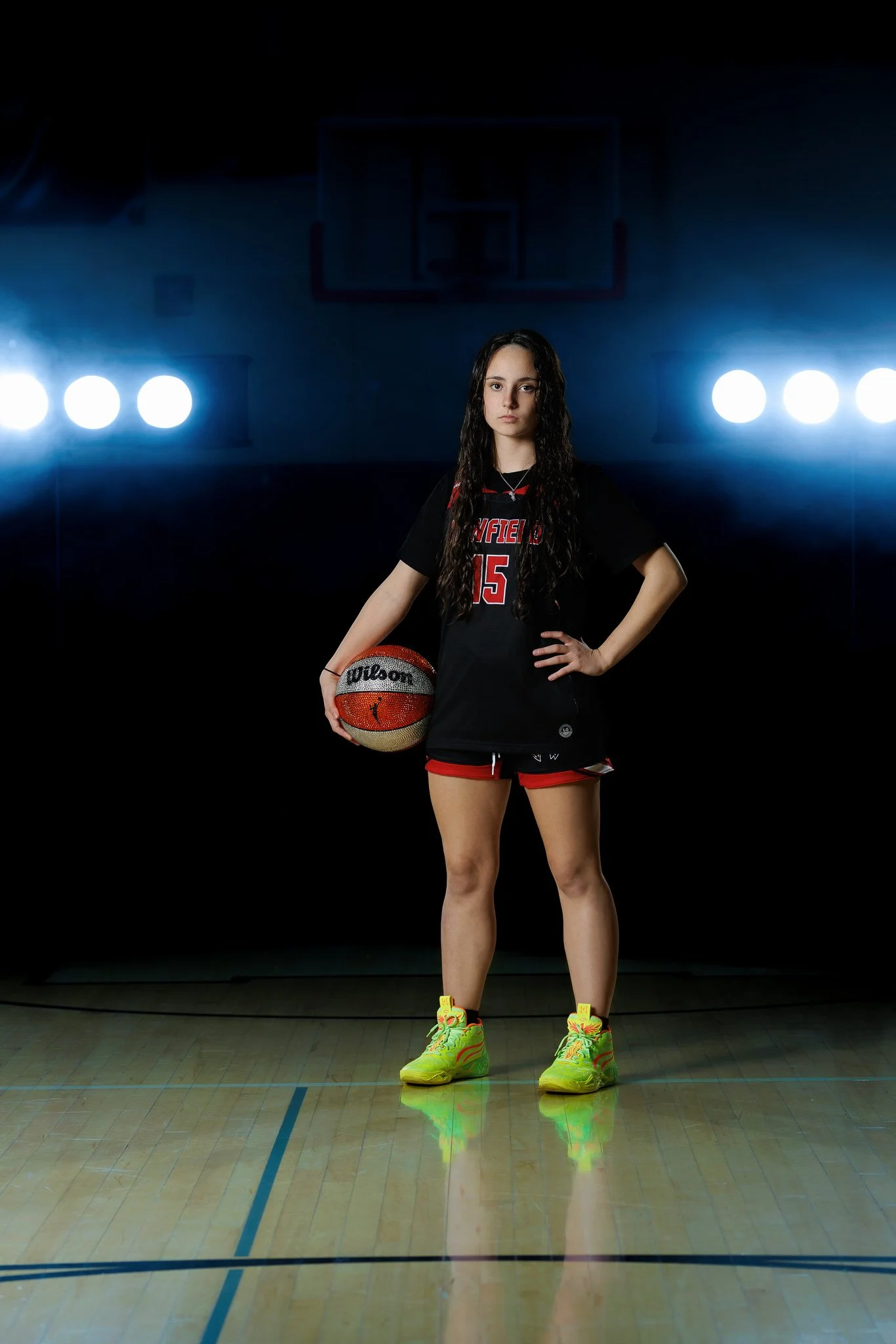 A young female basketball player standing on an indoor court, holding a basketball, with bright lights behind her.