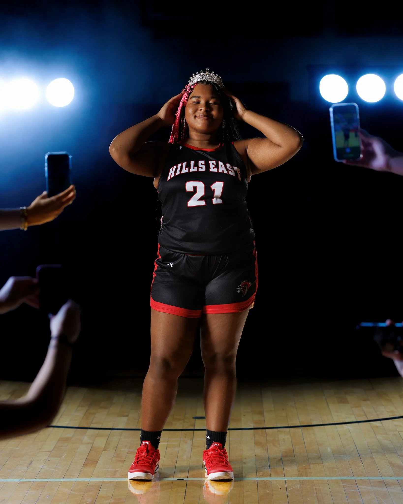 A young woman in a basketball uniform with the number 21 and 'Hills East' written on it, standing on a basketball court, wearing a tiara, surrounded by people taking photos.