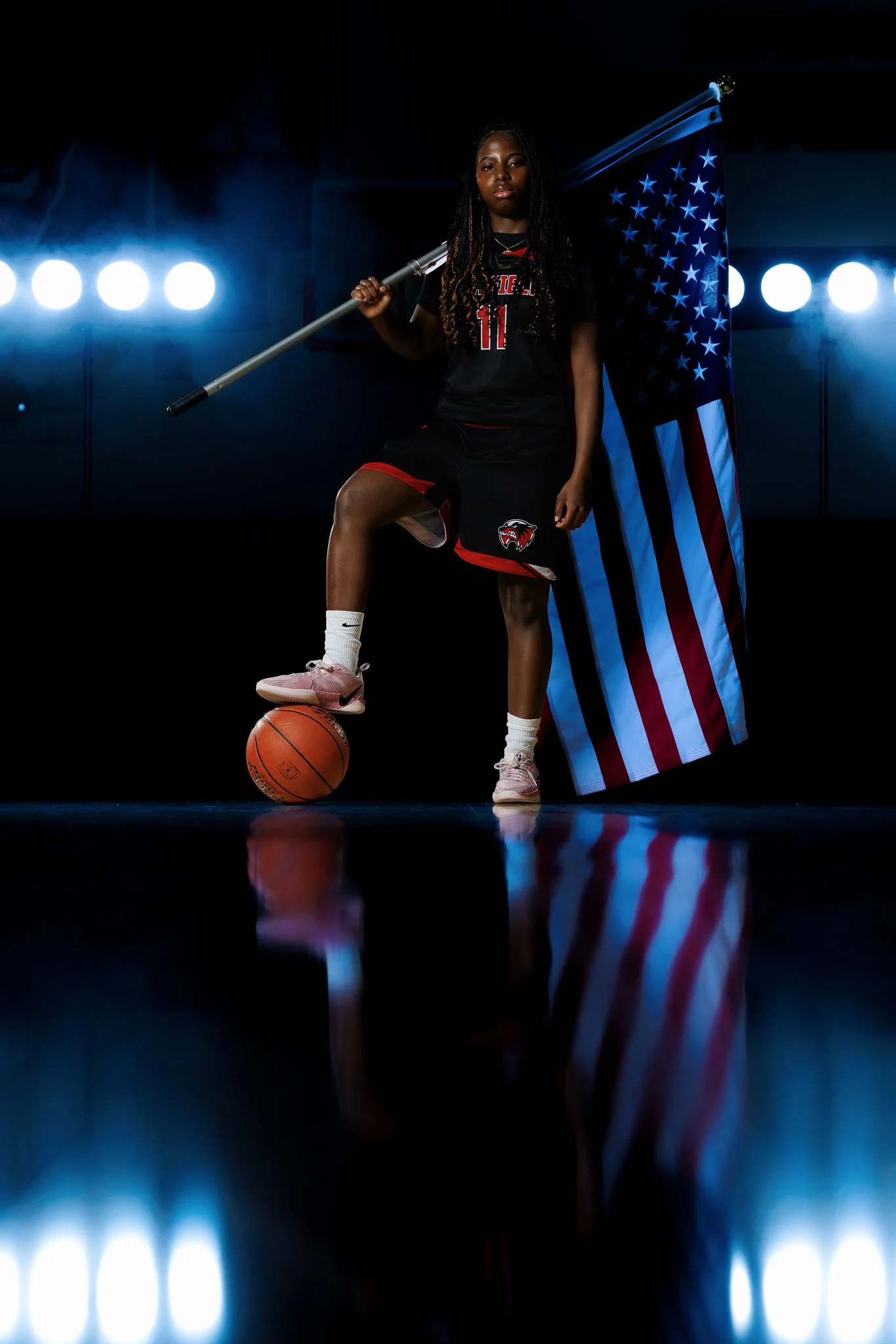 A female basketball player in a black uniform with red details holding a United States flag over her shoulder, standing on a basketball with her foot, in a dimly lit studio with bright lights behind her.
