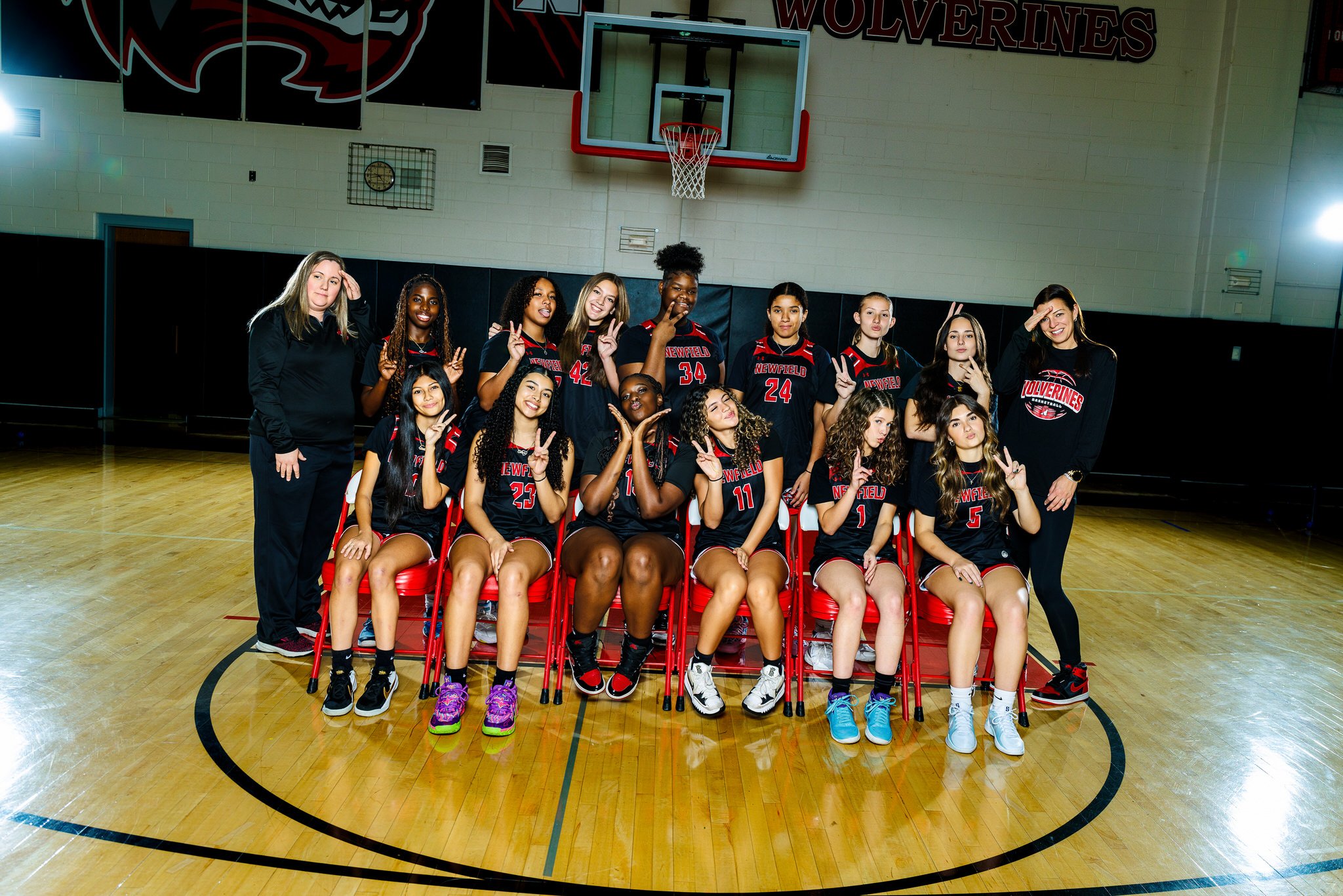 A girls' basketball team in black uniforms sitting and standing in a gymnasium, with two coaches on either side, posing for a team photo.