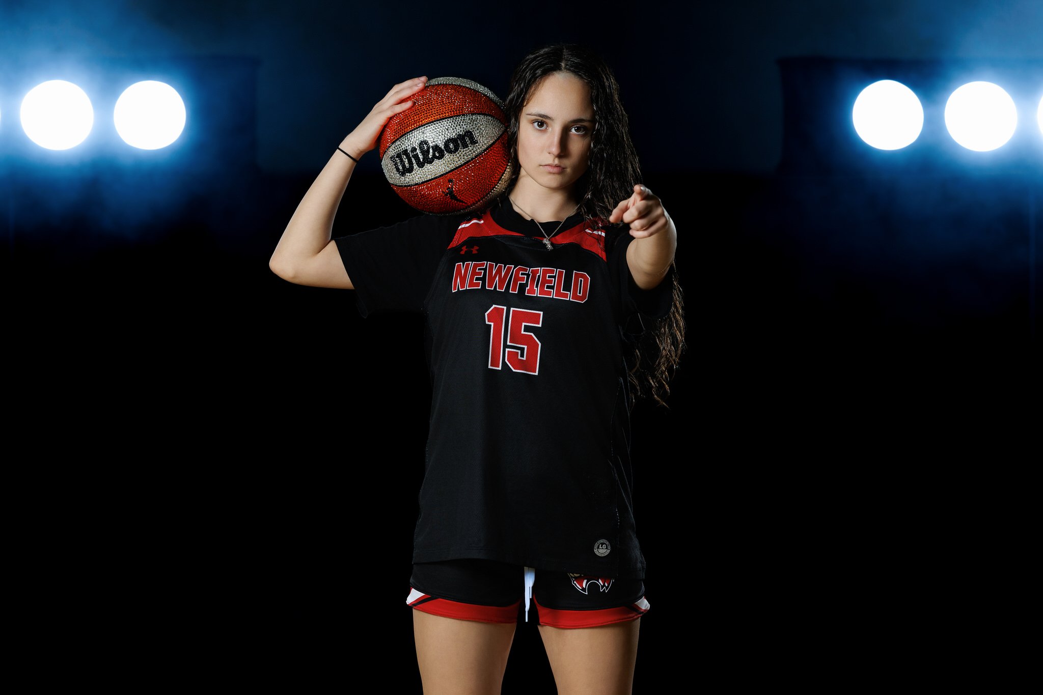 A young female basketball player stands in a dark court with bright lights behind her, holding a red and black Wilson basketball on her shoulder, pointing directly at the camera, wearing a black and red Newfield jersey and shorts.