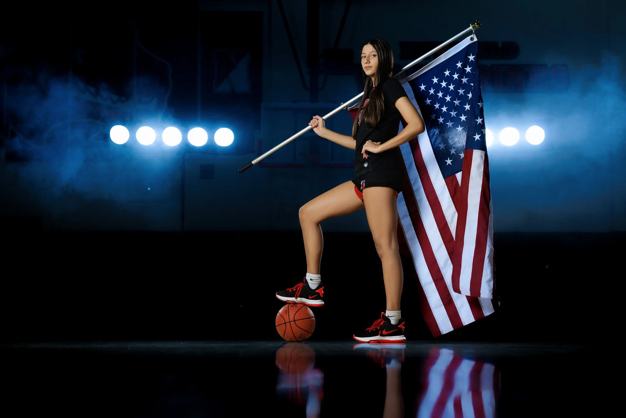 Young woman holding an American flag, standing on a basketball court with one foot on a basketball, wearing athletic shorts, a black t-shirt, and Nike sneakers, with a basketball court and bright lights in the background.