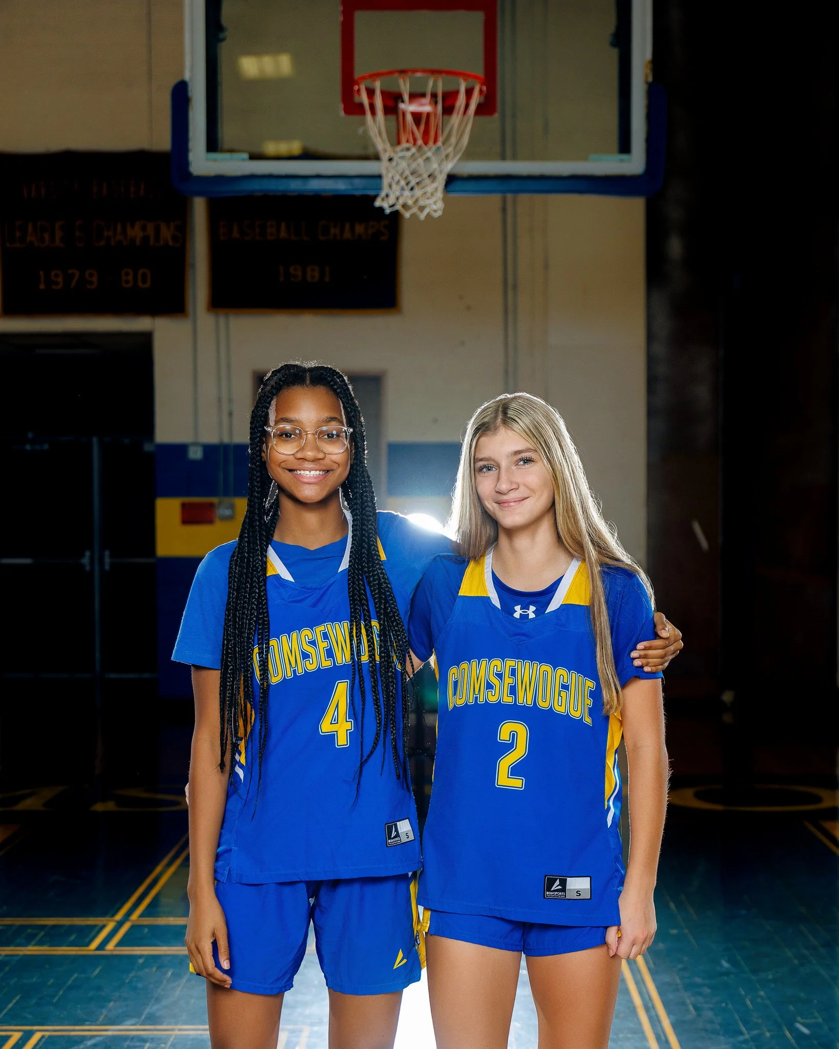 Two young female basketball players in blue and yellow uniforms standing in a gymnasium with a basketball hoop above them, smiling and posing for the photo.