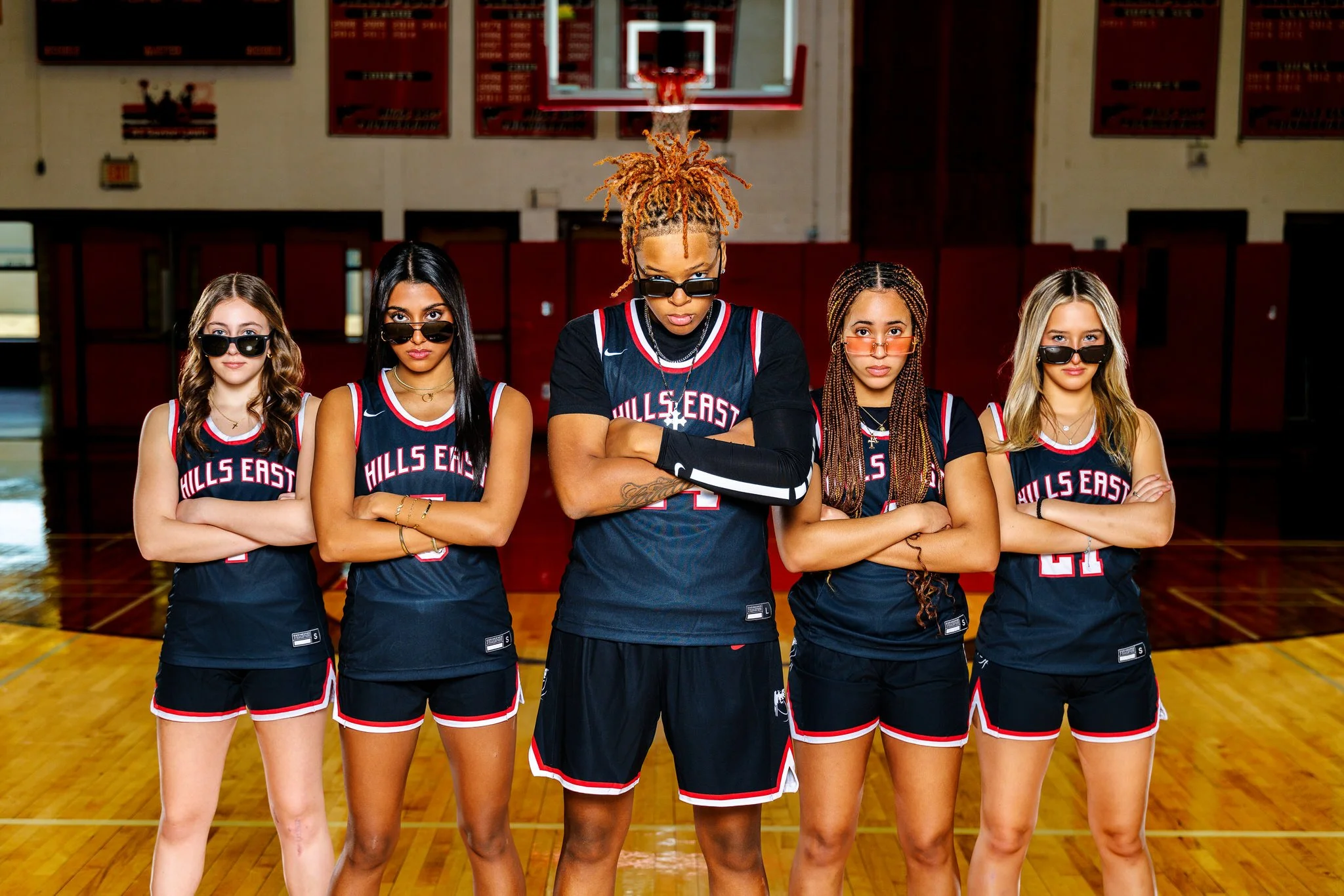 Five young women in basketball uniforms standing in a gymnasium with arms crossed, wearing sunglasses, with a basketball hoop in the background.