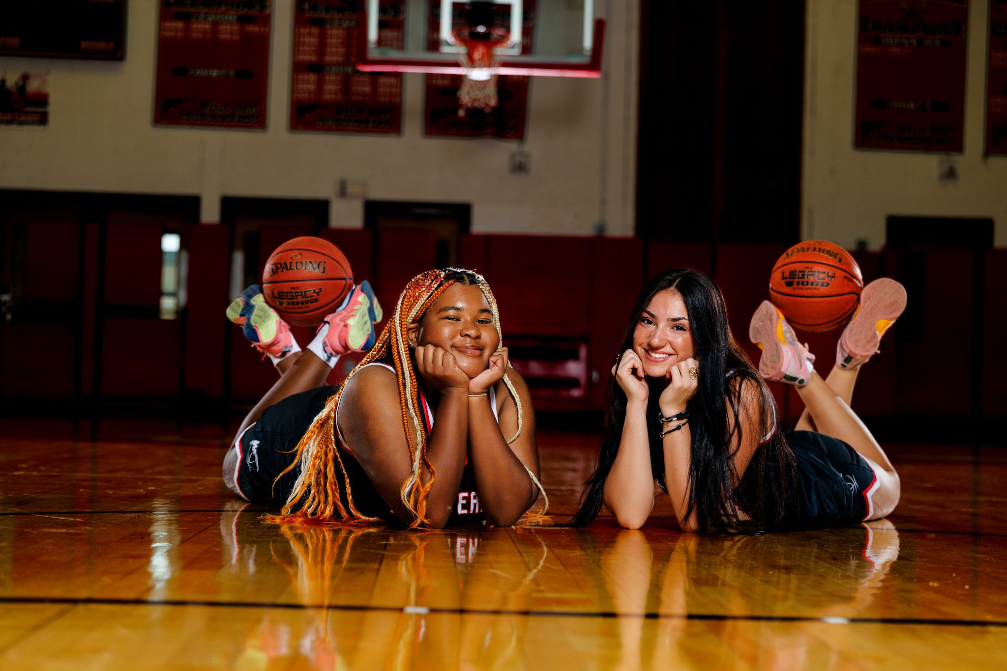 Two young women lying on a basketball court with their heads resting on their hands, smiling and looking at the camera, with basketballs behind them and a basketball hoop overhead.