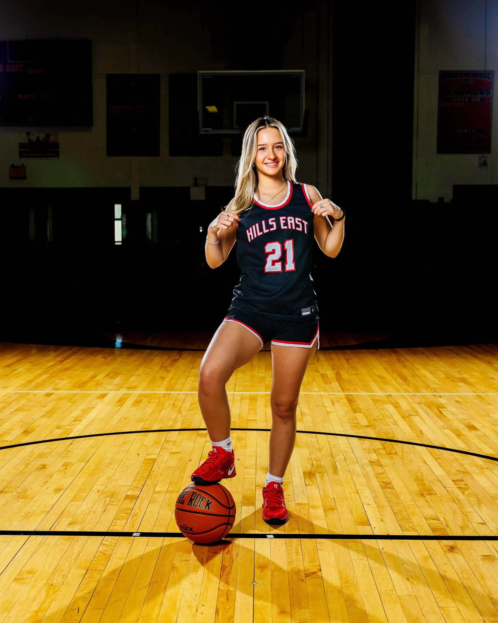 Young woman in a basketball uniform with the number 21 and 'Hills East' on it, standing on a basketball court with a basketball under her foot, smiling at the camera.