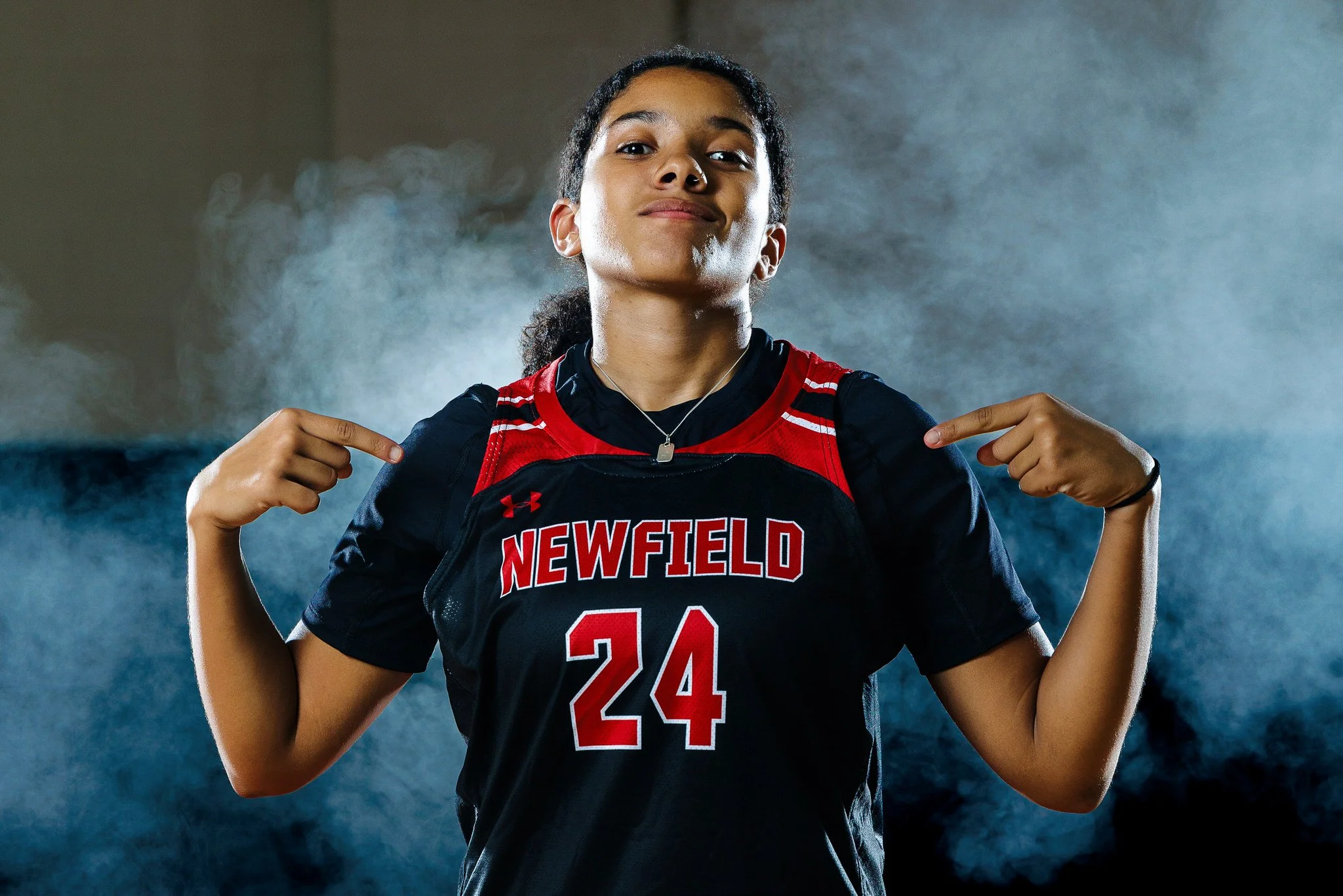 A young female athlete in a black and red sports jersey with the word 'Newfield' and the number 24, pointing to her chest with both hands, standing in a smoky, well-lit environment.