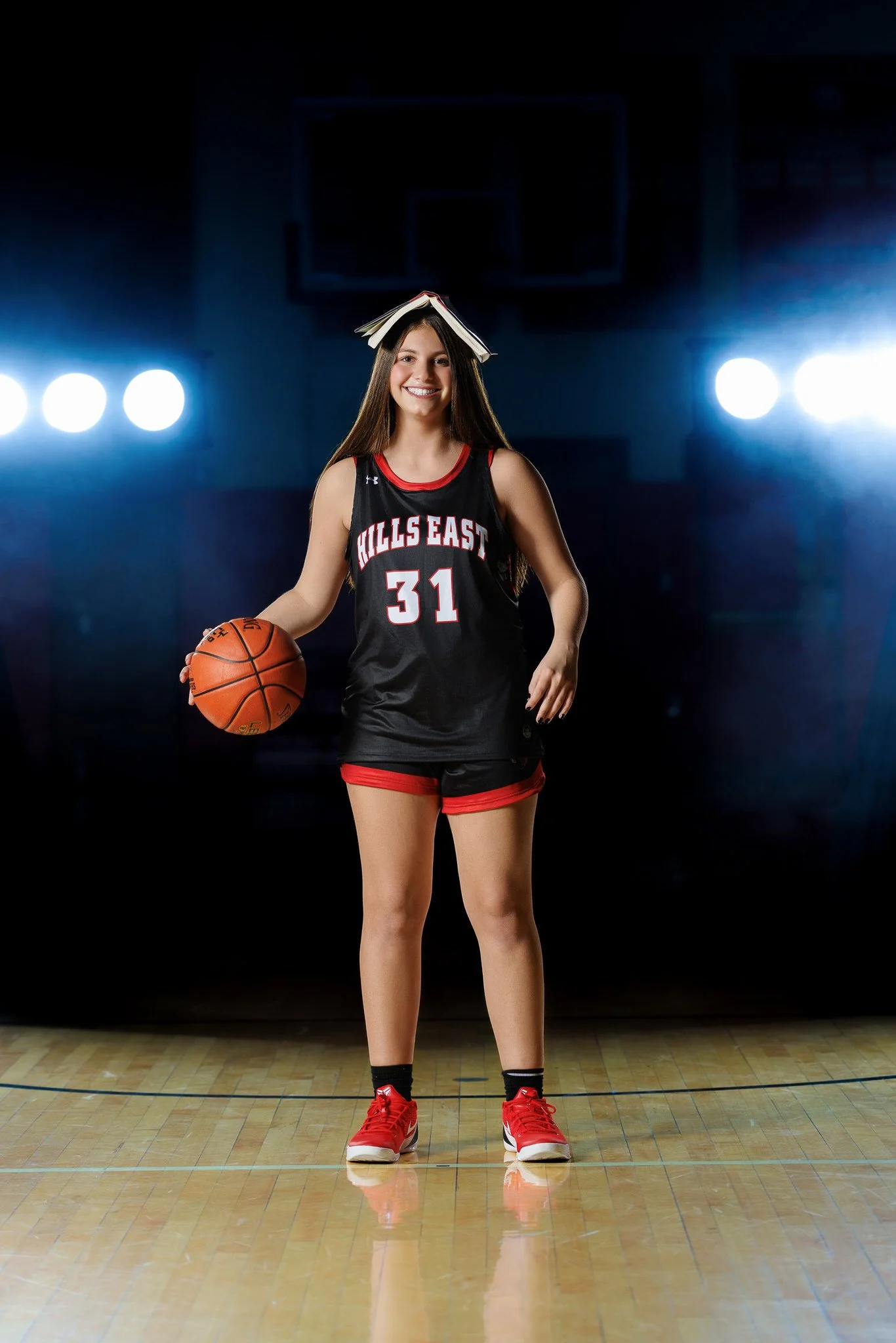 A young woman in a black and red basketball uniform with the number 31, holding a basketball, standing on a gym court with bright lights behind her, with a book balanced on her head and smiling.