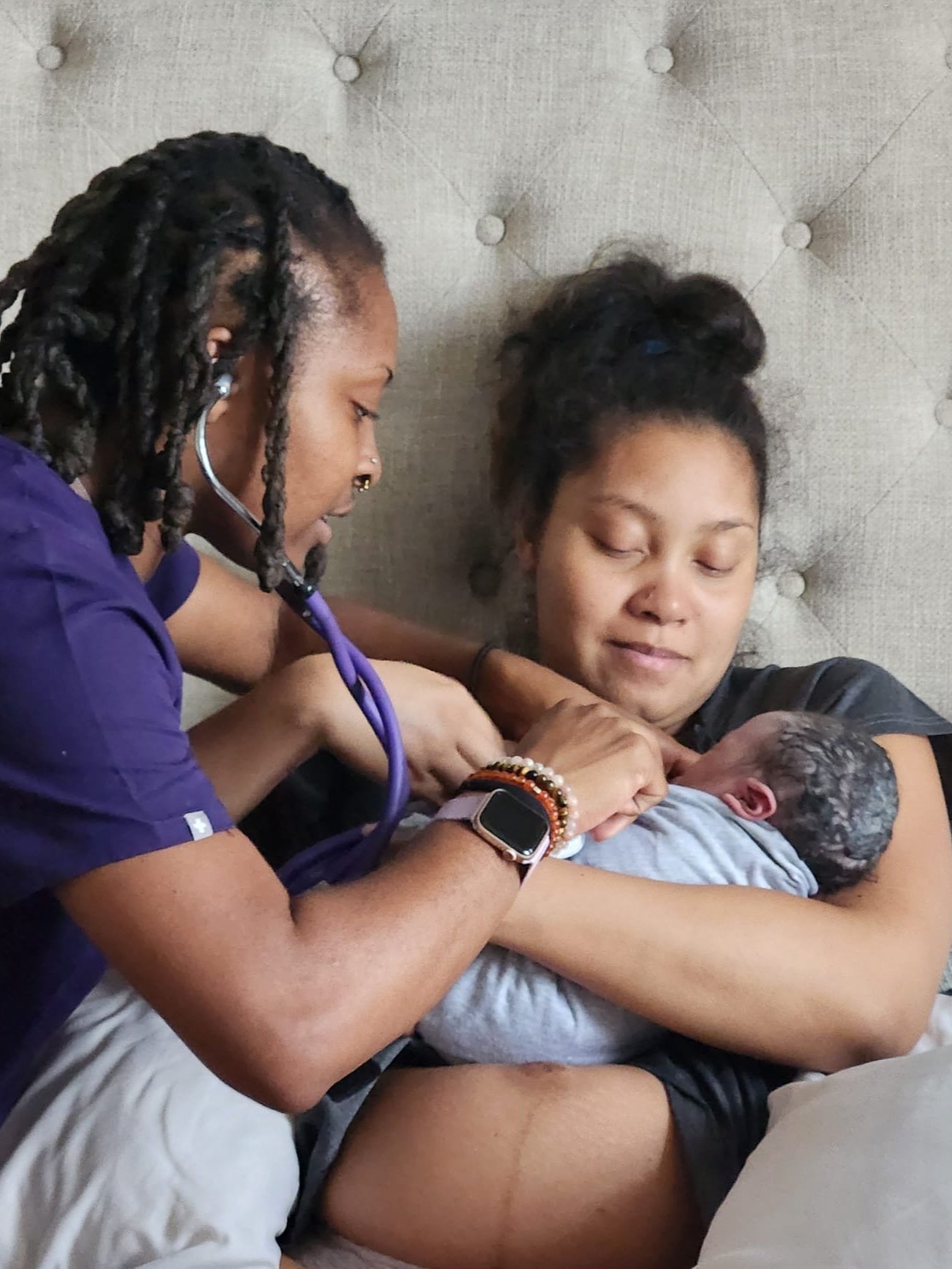 A new mother holding her newborn in a blanket, while a midwife or nurse checks the baby using a stethoscope.