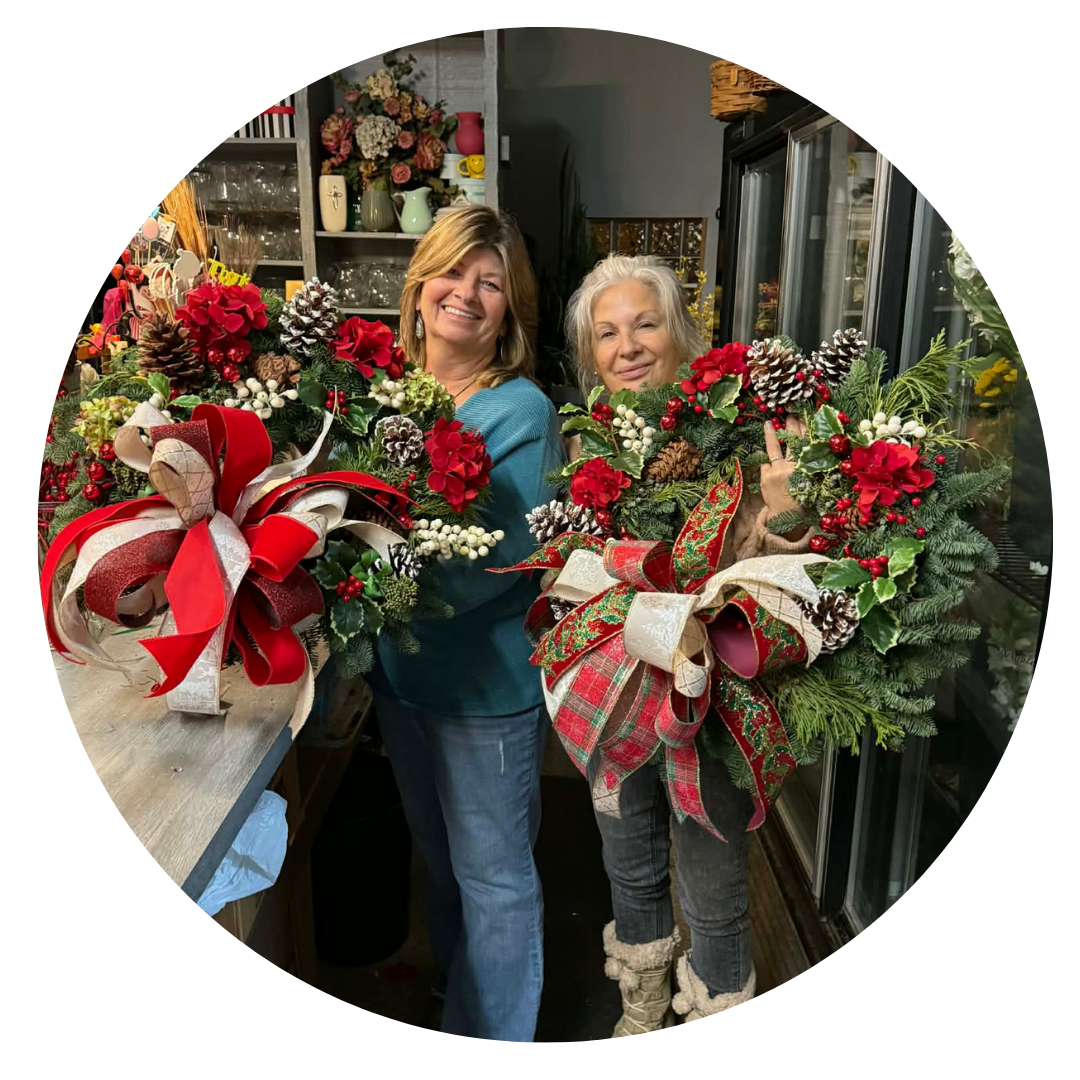 Two women standing indoors, each holding a large Christmas wreath decorated with bows, pinecones, and red and white accents. The woman on the left is smiling, wearing a teal sweater, while the woman on the right is making a gesture with her middle finger, wearing a tan outfit and boots.