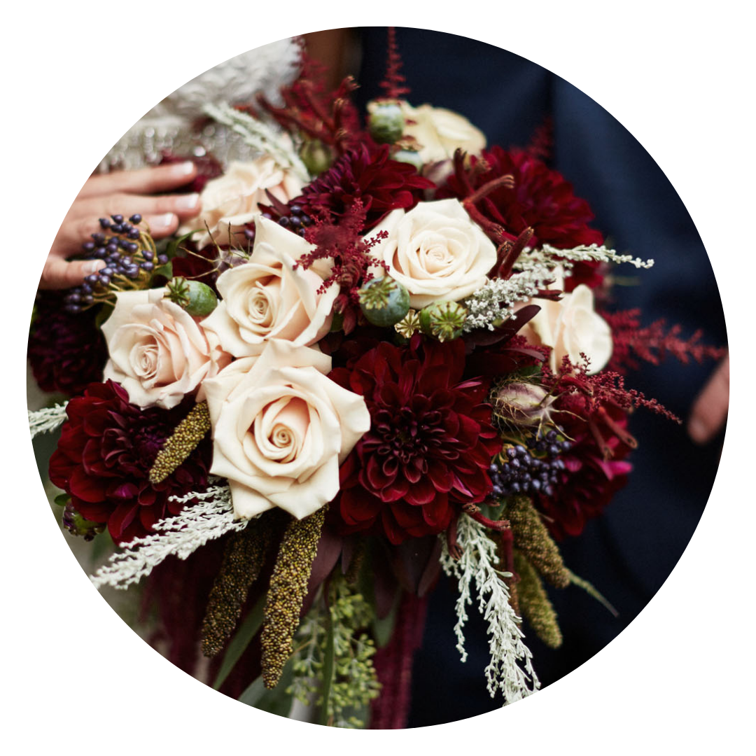 A person holding a bouquet of cream-colored roses, deep red dahlias, purple berries, and greenery, with a dark background.
