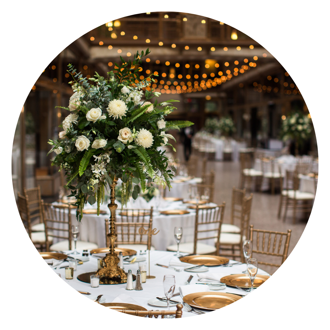 Elegant banquet table decorated with a tall floral centerpiece featuring white flowers and green foliage, set in a dimly lit venue with string lights overhead.