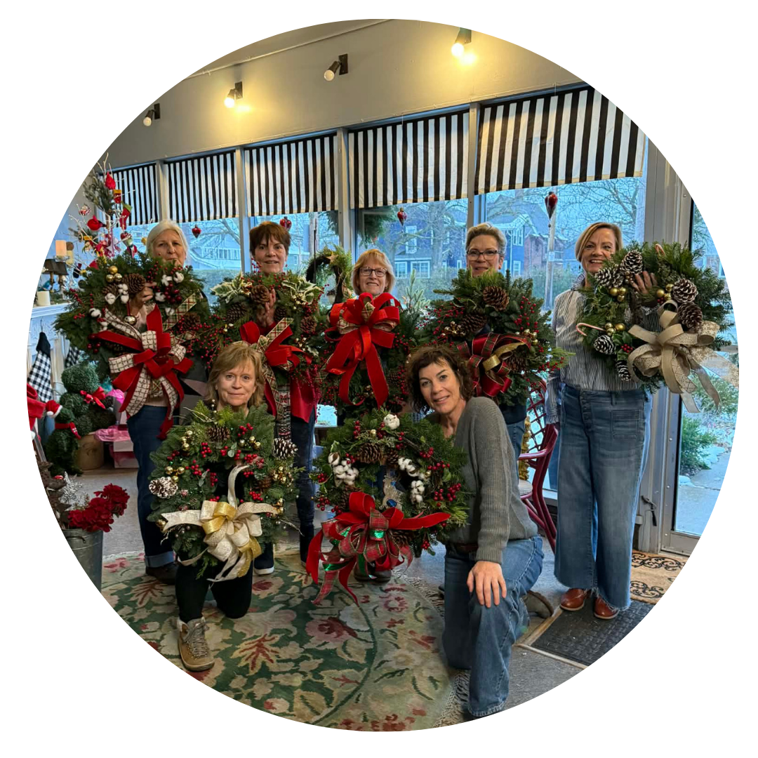 Group of women holding decorated Christmas wreaths inside a room with large windows and holiday decorations.