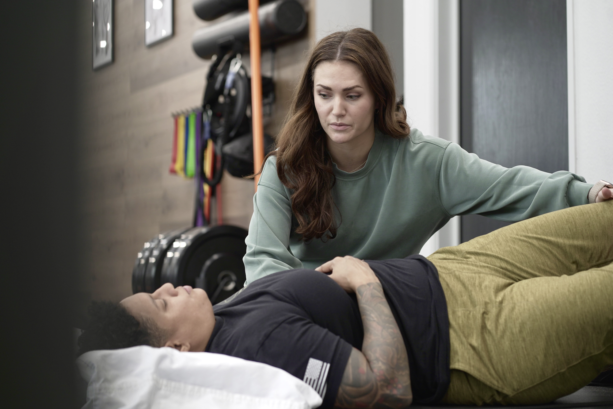 A physical therapist attending to a patient in a gym setting, with exercise equipment like weights and resistance bands in the background.