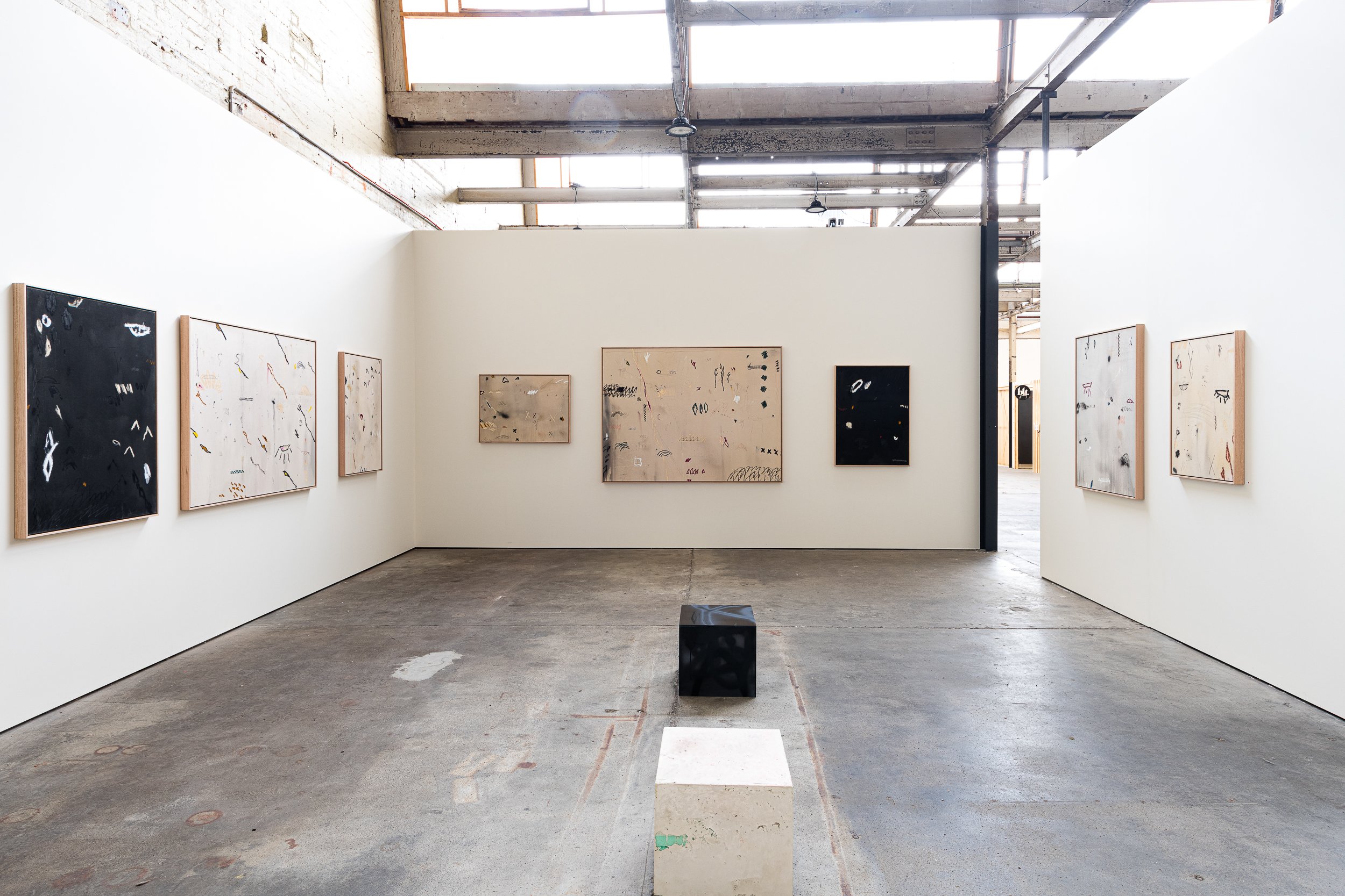 Art gallery interior with abstract paintings on white walls and concrete floor, featuring scattered minimalist artworks in frames and two cube-shaped floor stools.