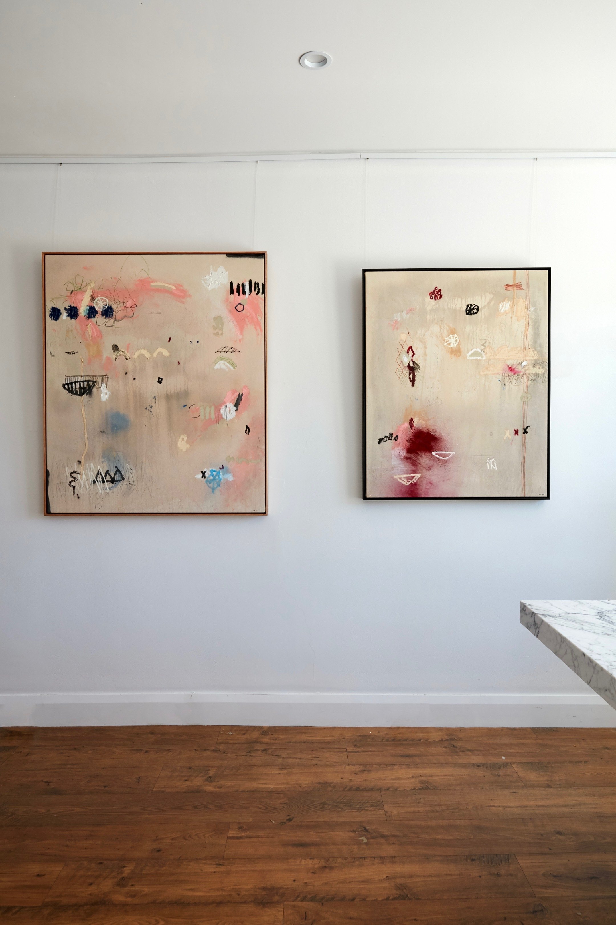 Two abstract paintings hanging on a white wall above a wooden floor, featuring various shapes and colors such as pink, black, and blue.