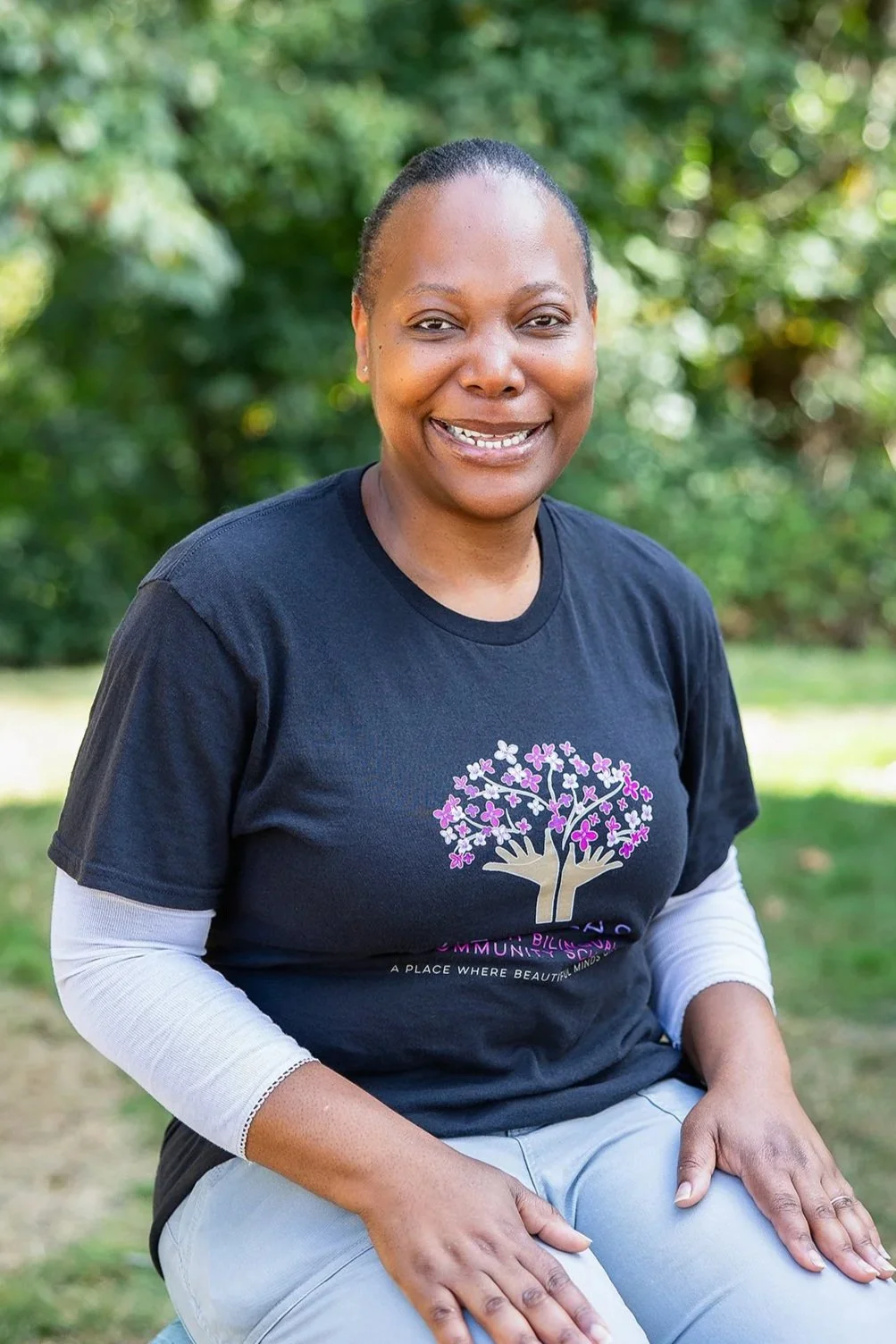 Une femme noire souriante, vêtue d'un t-shirt orné d'un motif d'arbre, assise en plein air sur fond de verdure feuillue