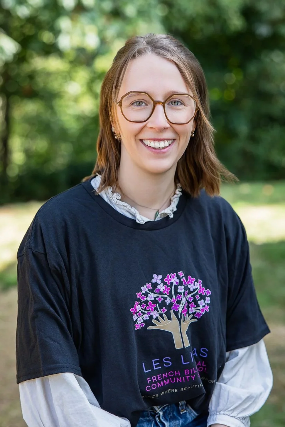 Femme souriante portant des lunettes et un t-shirt de la communauté bilingue française « Les Lilas », à l'extérieur