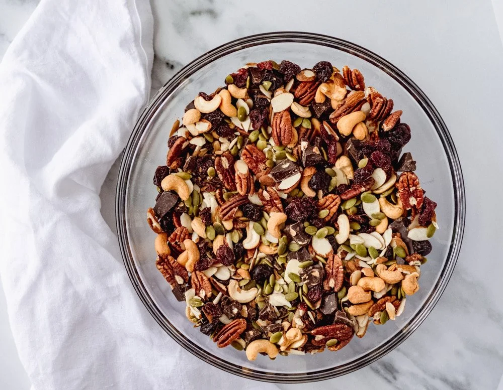 A glass bowl filled with trail mix containing nuts, seeds, dried fruit, and dark chocolate chunks on a marble surface with a white cloth.