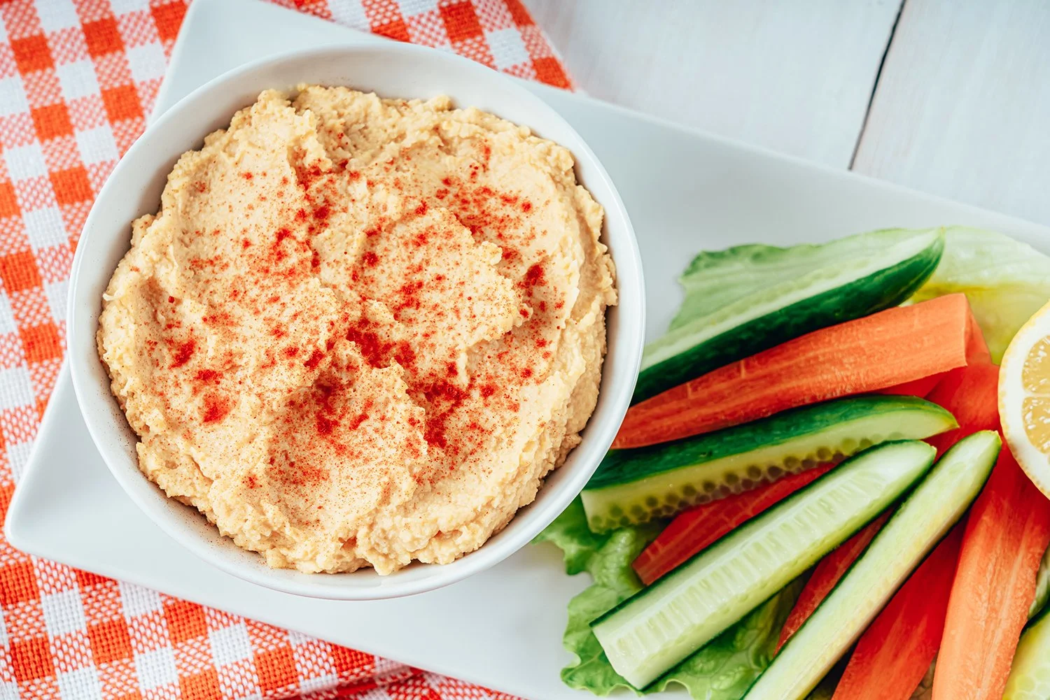 A bowl of hummus sprinkled with paprika is served on a plate with fresh cucumber, carrot sticks, lettuce, and a lemon wedge.