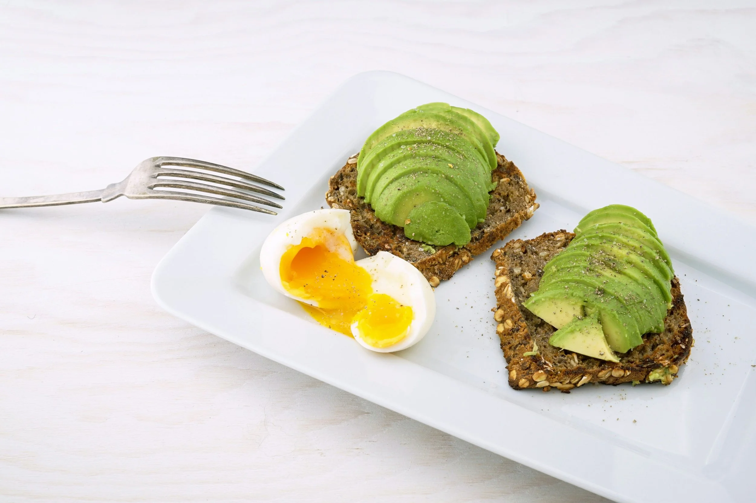 Two slices of seeded toast topped with avocado and a soft-boiled egg with a runny yolk on a white plate.