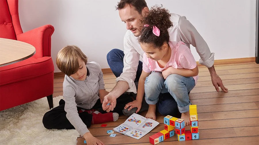 Father playing and learning with two children using colorful letter blocks and activity books.