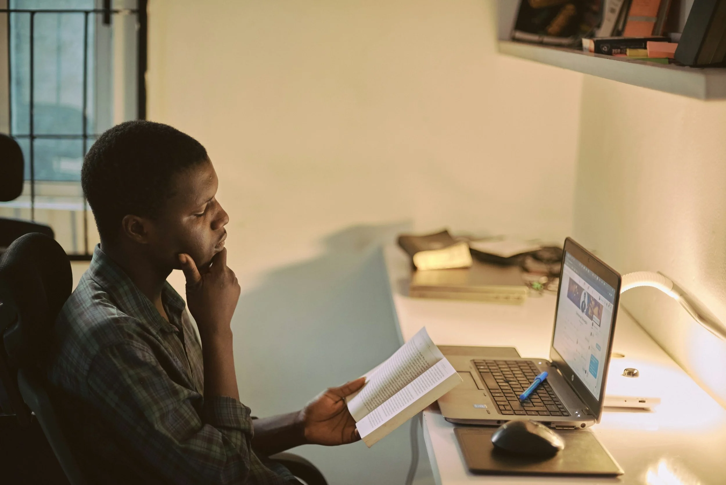 A young man sits at a desk studying, holding a book in one hand and thoughtfully looking at a laptop screen.