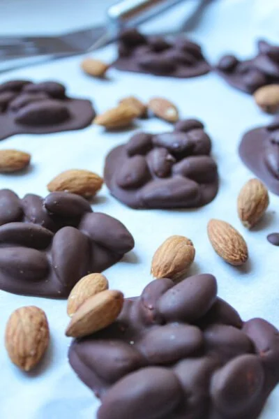 Close-up of clusters of almonds coated in dark chocolate, arranged on parchment paper with a few loose almonds scattered around.